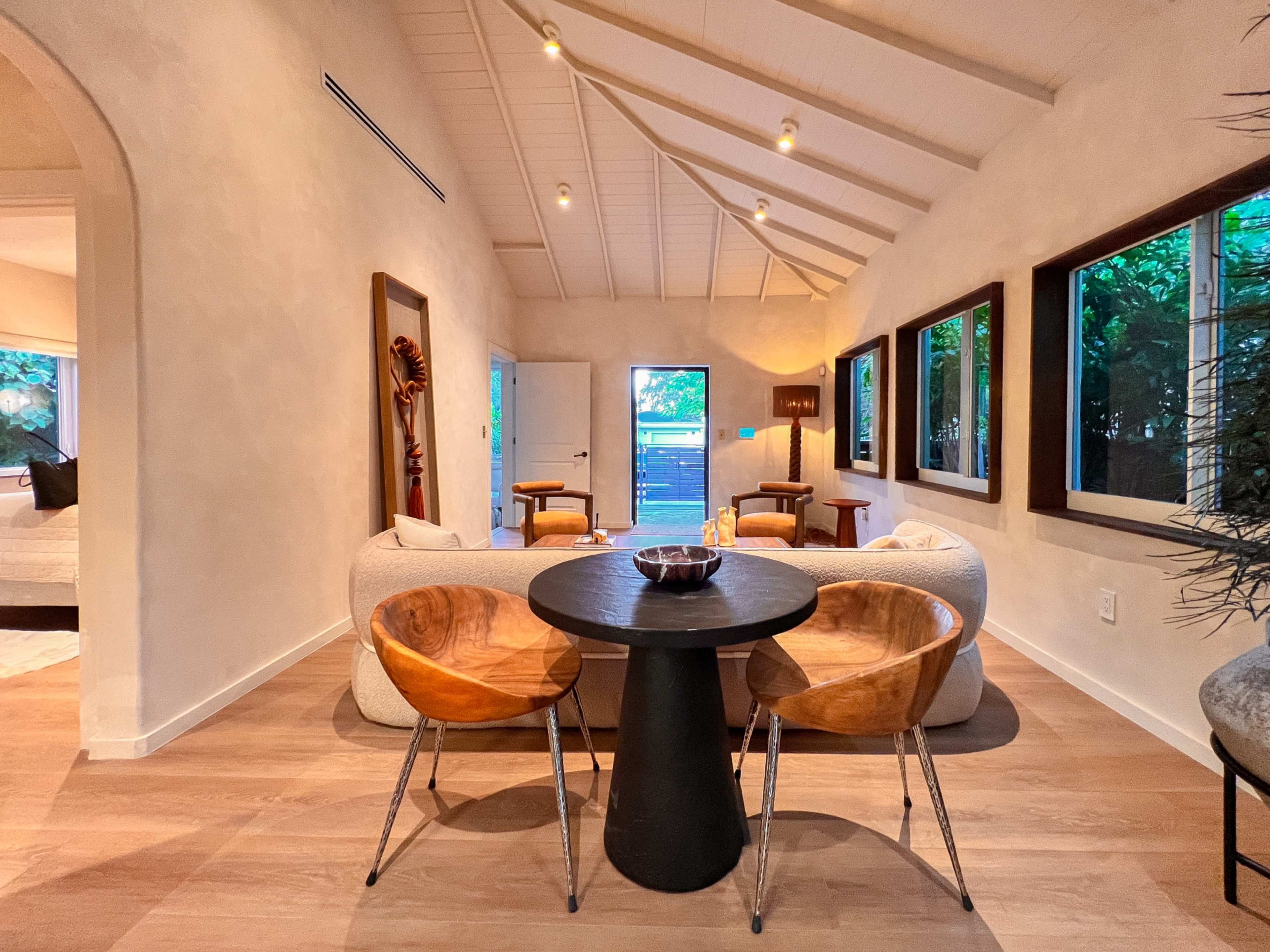 A cozy interior features a circular black table surrounded by wooden chairs, with a seating area and large windows allowing natural light.