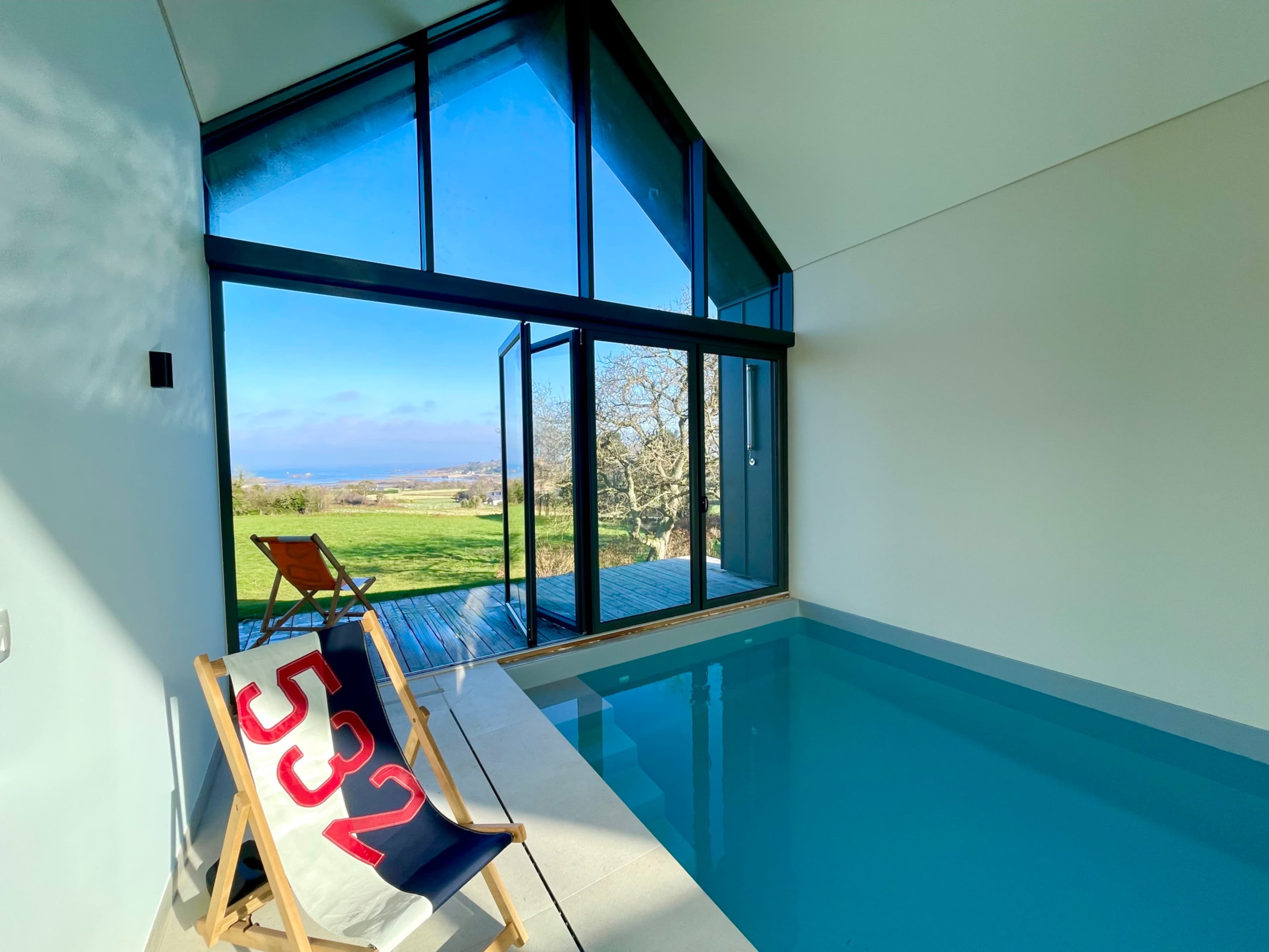 A modern indoor pool area features large glass doors opening to a scenic landscape with a view of trees and the ocean.