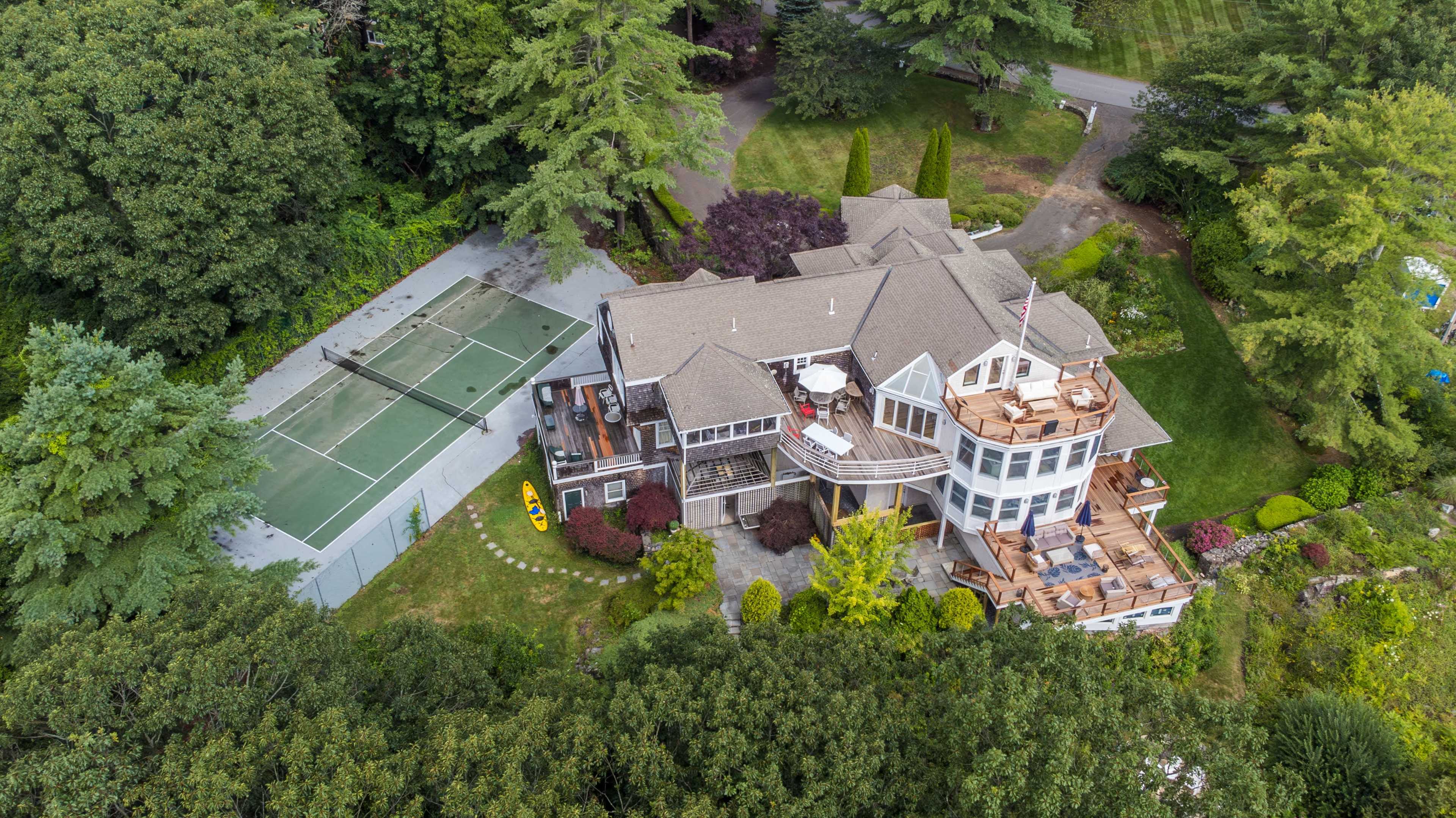 A large, multi-level house with a surrounding green lawn, a wooden deck, and a nearby tennis court.