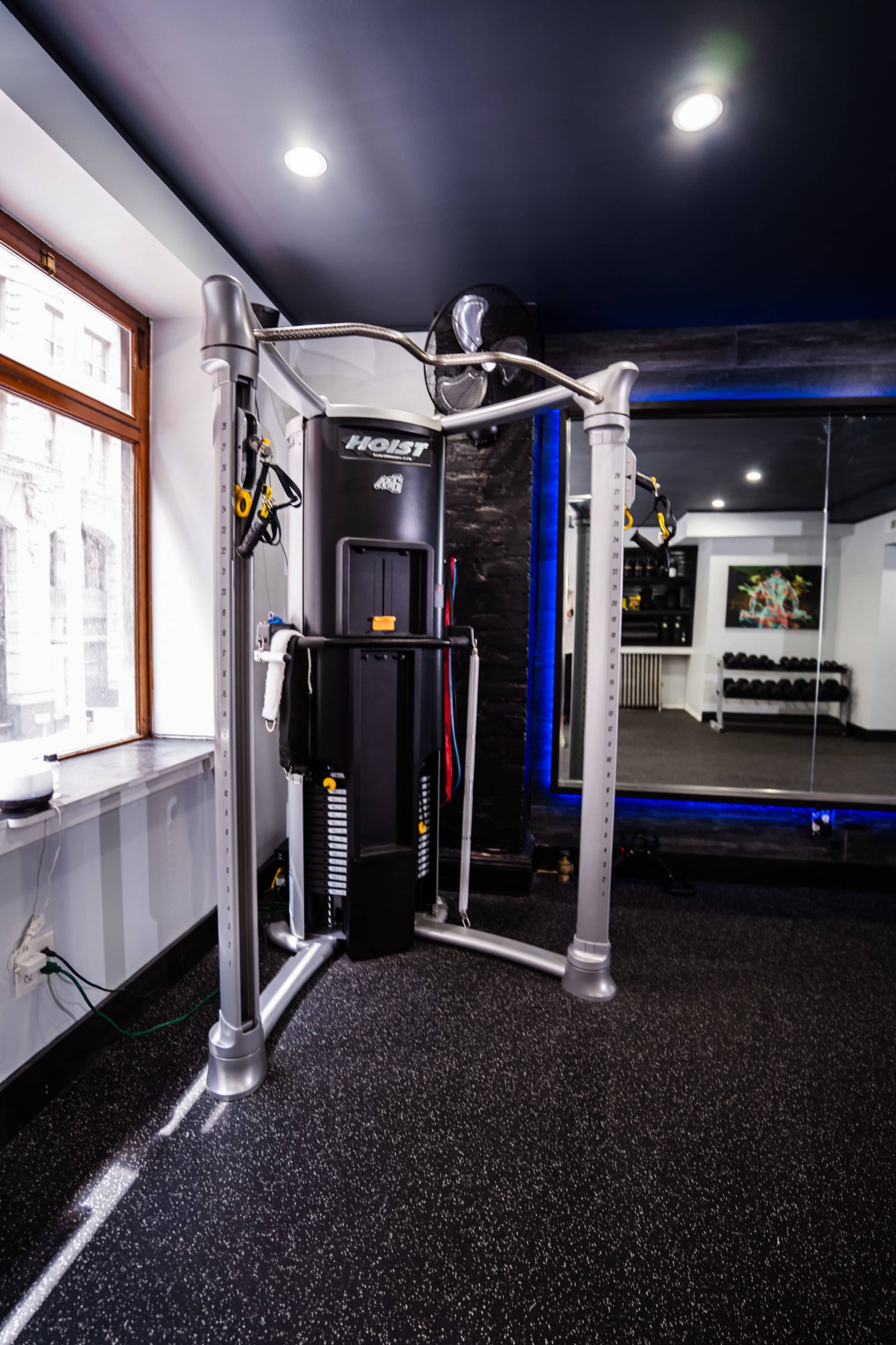A gym corner featuring a multi-functional weight machine against a wall, with a large mirror and free weights visible in the background.