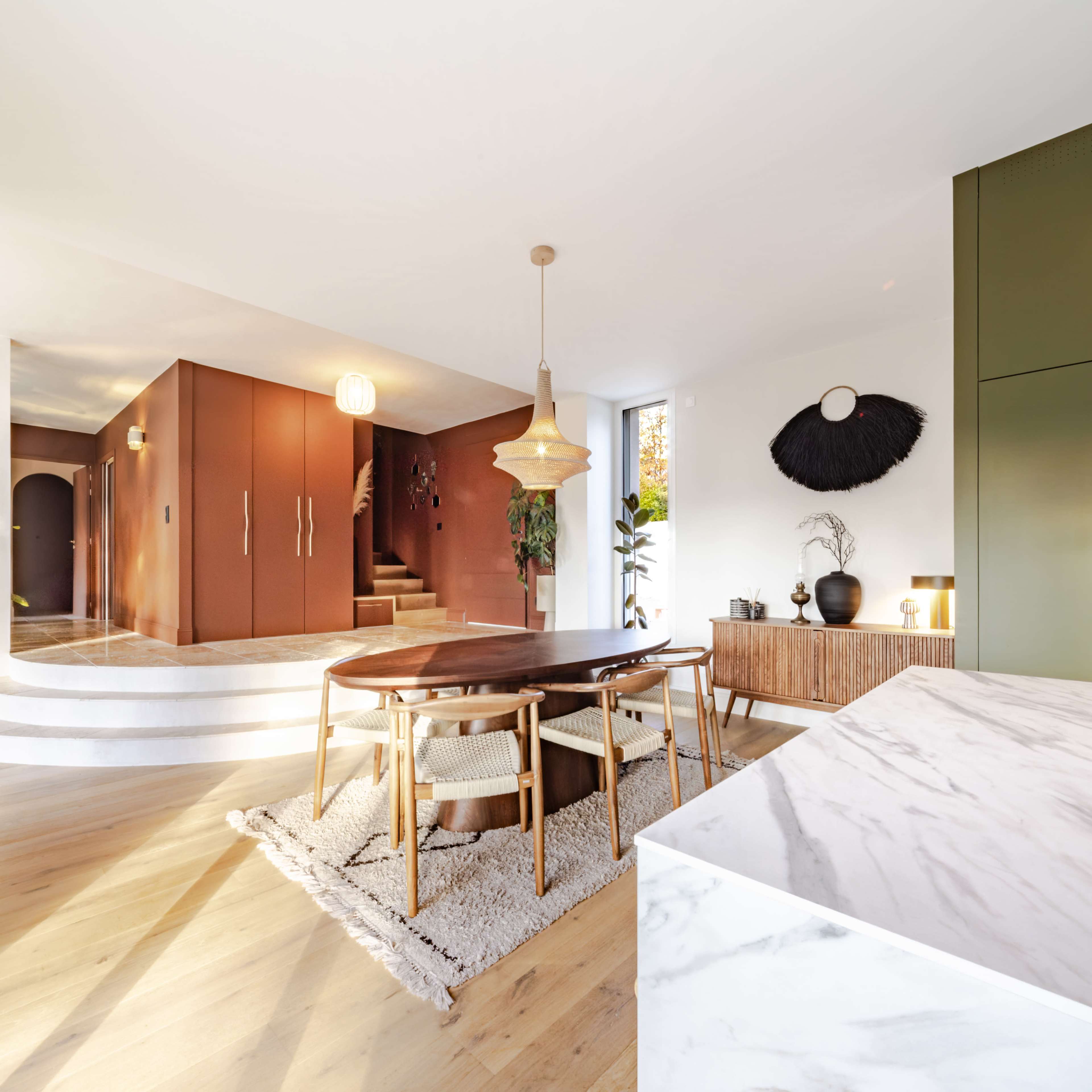 The image shows a modern dining area with a wooden table, four chairs, a patterned rug, and a kitchen island, all illuminated by natural light coming through large windows.
