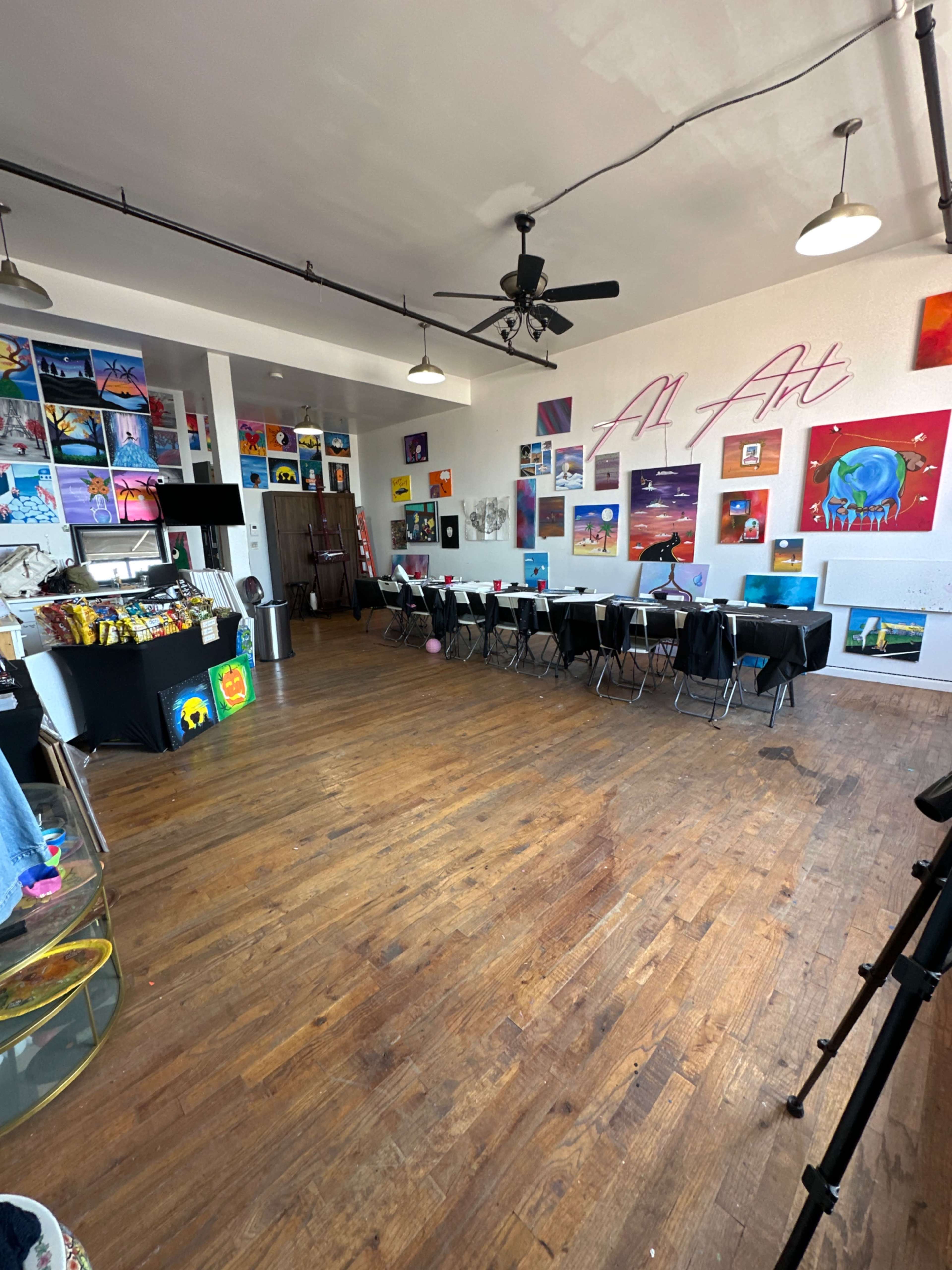An art studio with wooden floors, tables arranged in a row, and colorful paintings displayed on the walls.