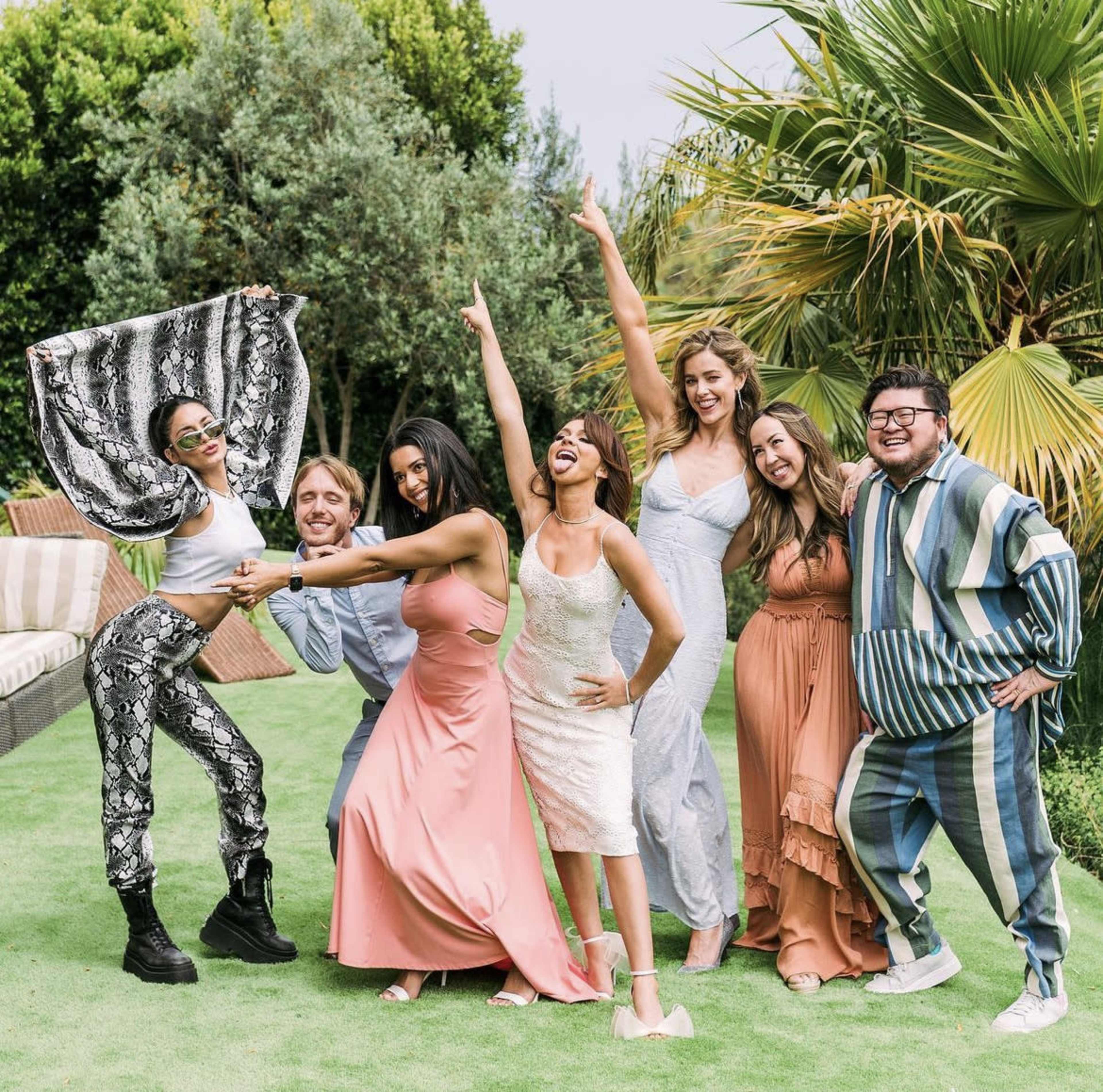 A group of seven people poses outdoors in a garden setting, wearing a variety of colorful outfits and striking playful poses.