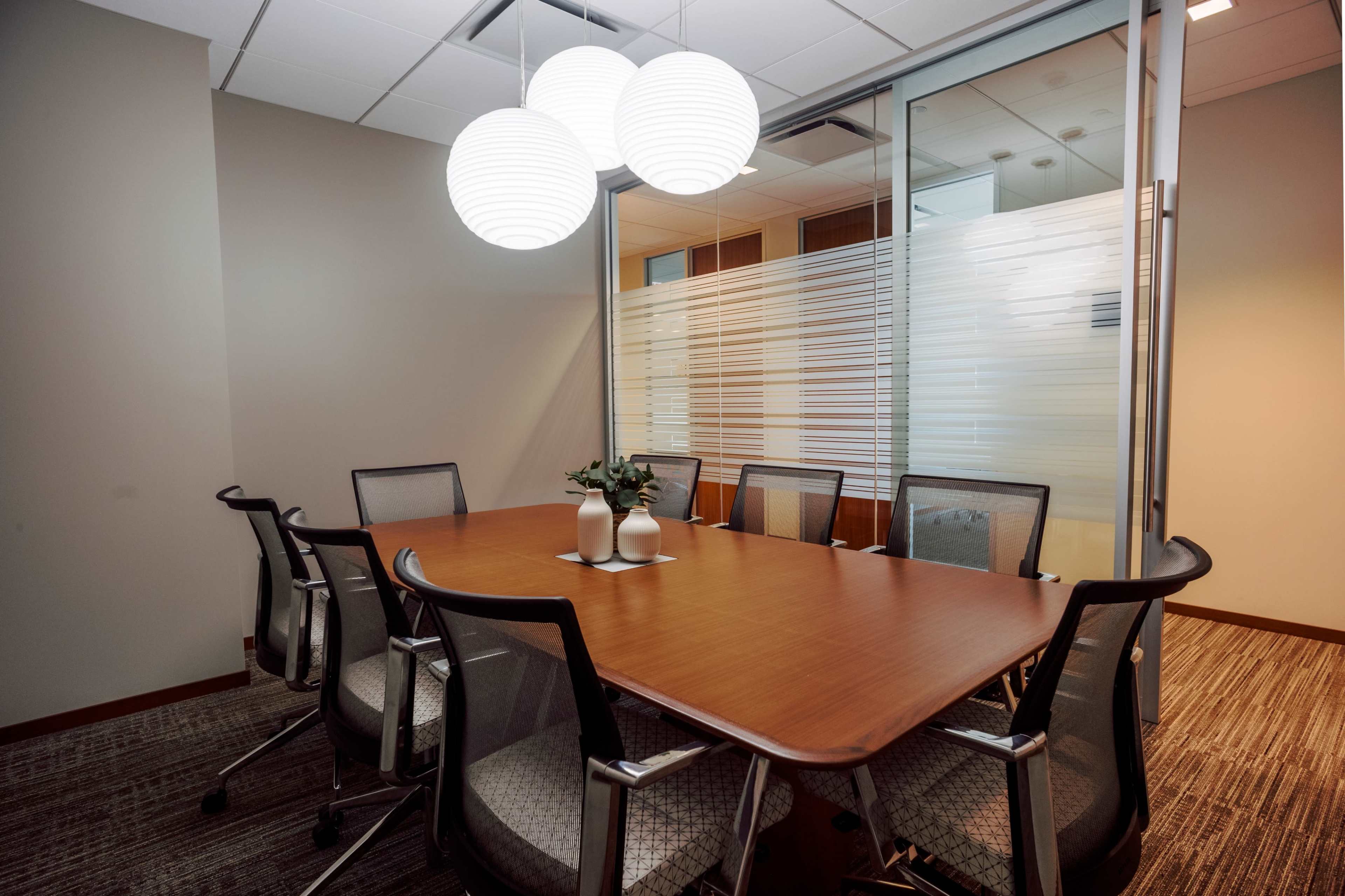 The image shows a modern conference room featuring a large wooden table surrounded by ergonomic chairs, with pendant lighting and a glass partitioned area.