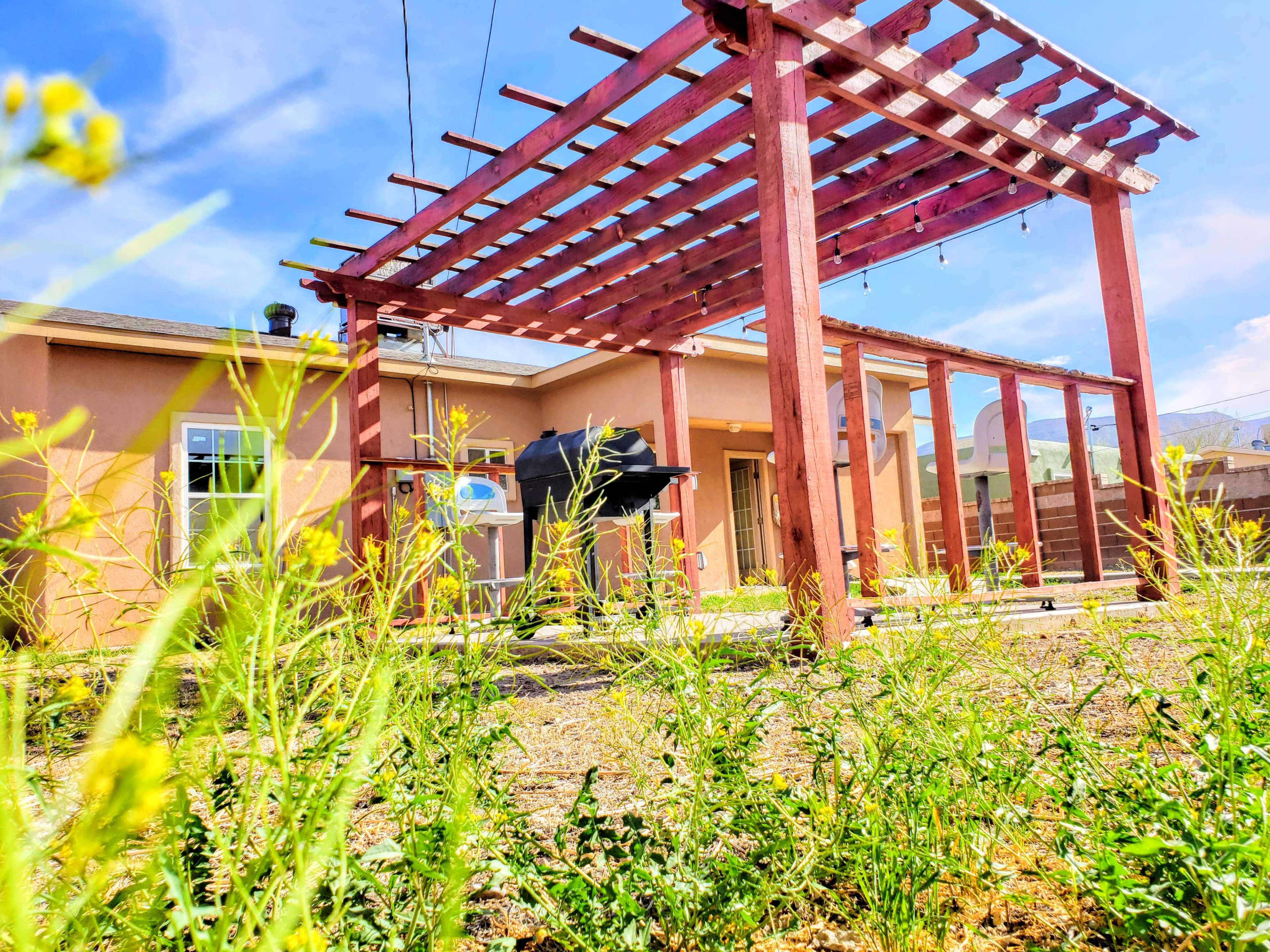 A wooden pergola stands over a grill in a backyard with wild plants growing nearby and a house in the background.