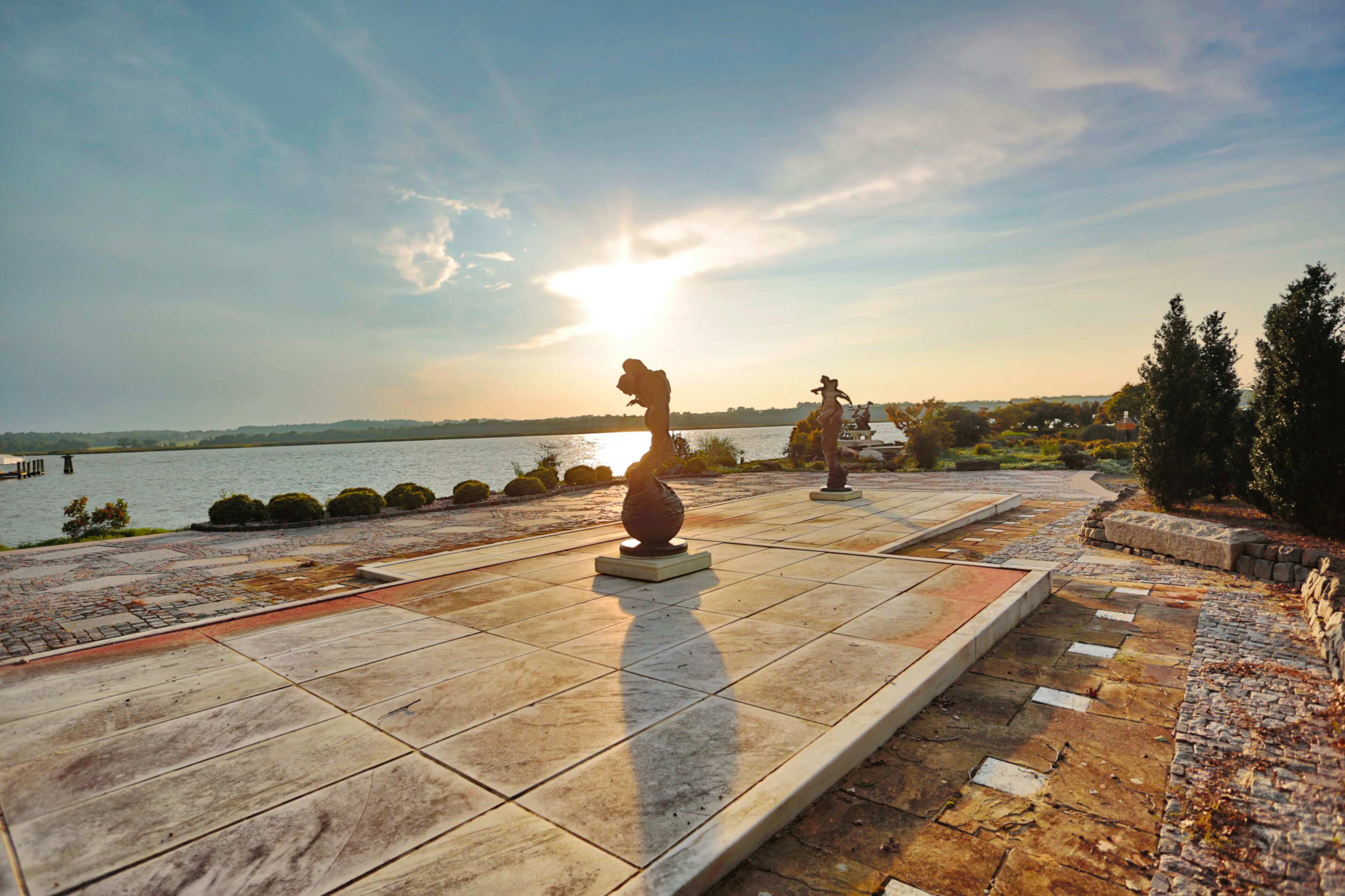 The scene shows a landscaped area with two sculptures positioned on a stone path, facing a river under a bright sky at sunset.