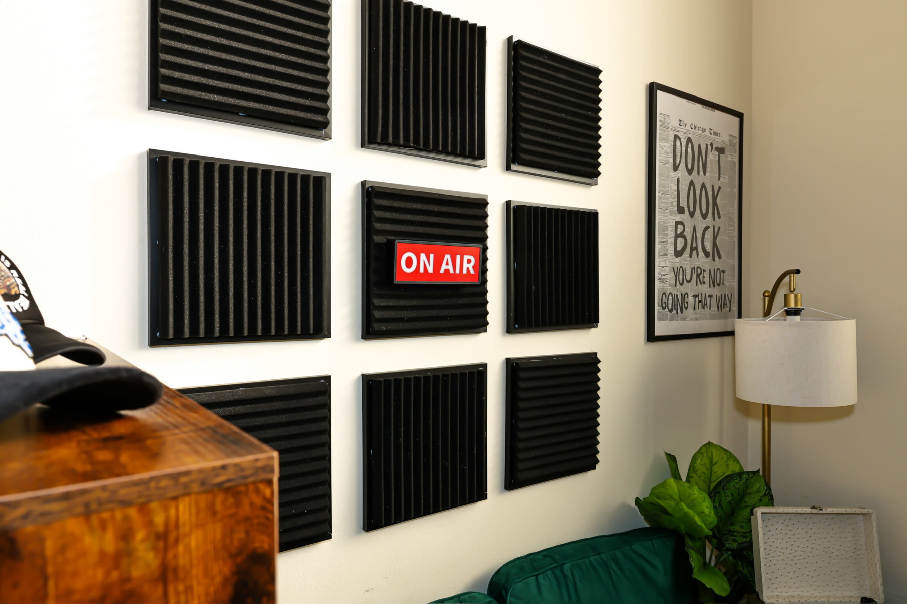 The image shows a wall with nine black acoustic panels and a red "ON AIR" sign, accompanied by a framed poster and a lamp near a green sofa.