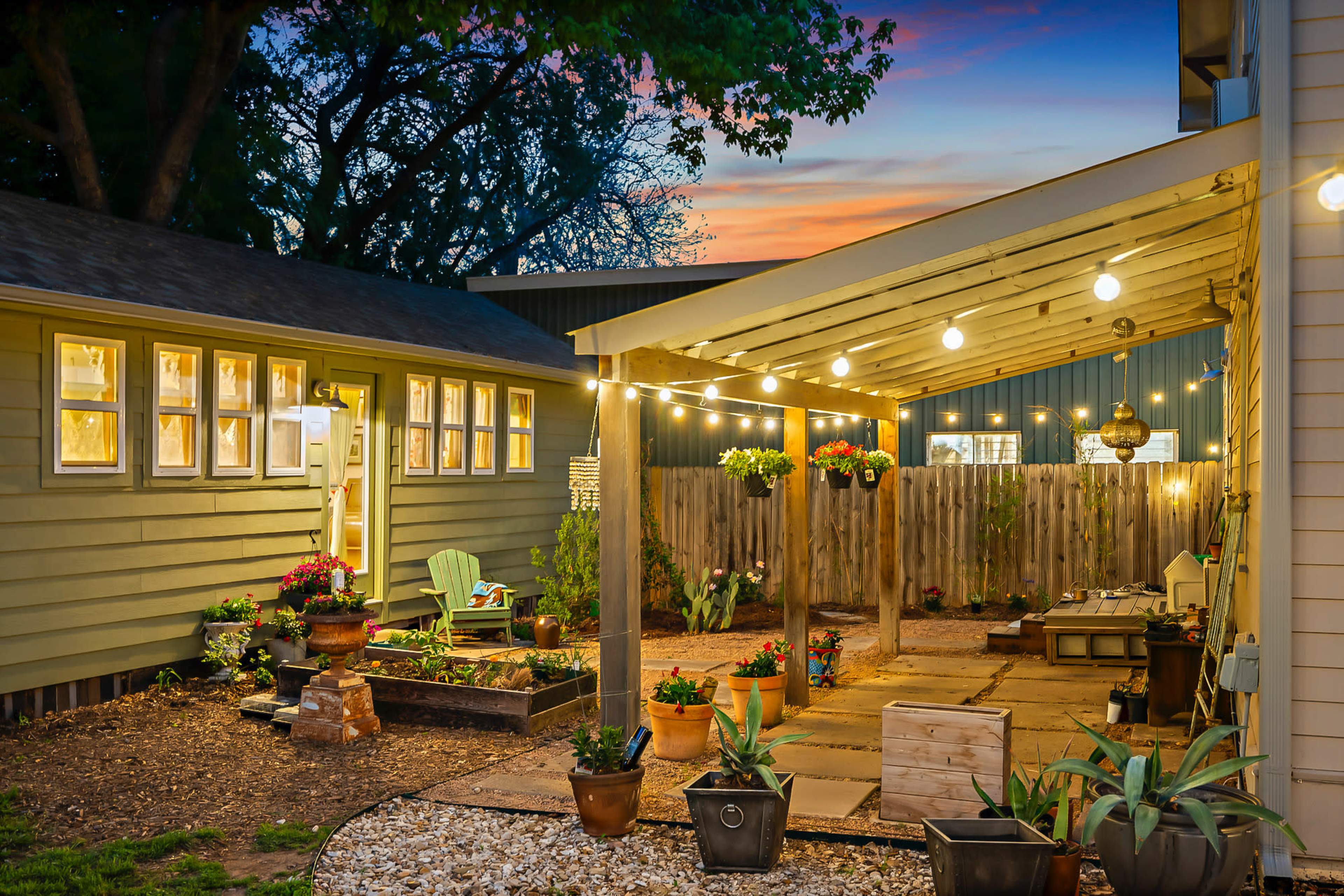 The image shows a well-lit patio area with string lights, surrounded by potted plants and a neighboring building, under a sunset sky.