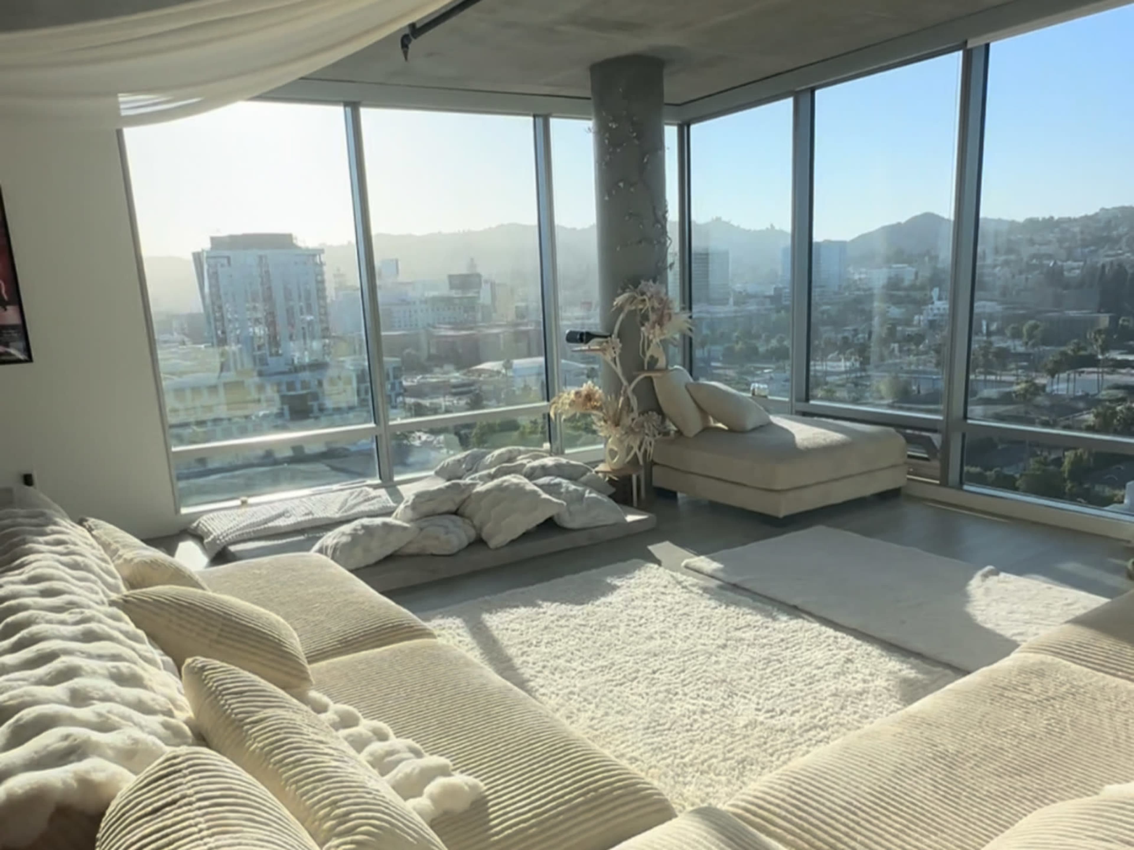 The image shows a spacious living room with large windows offering a view of a city skyline, furnished with a plush sofa and a light-colored area rug.