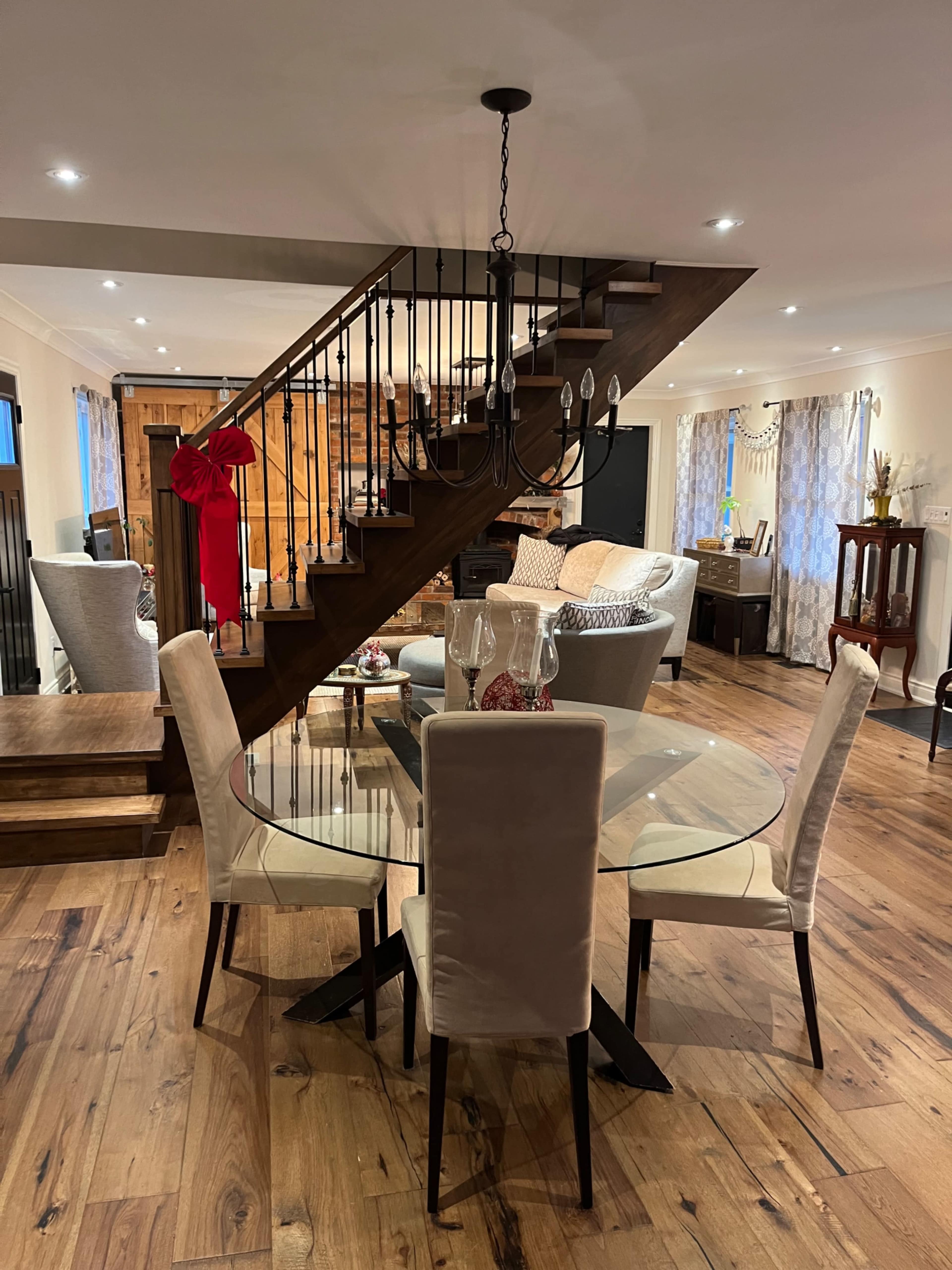 A glass dining table with four chairs sits in a room featuring a staircase with wooden steps and a chandelier above.