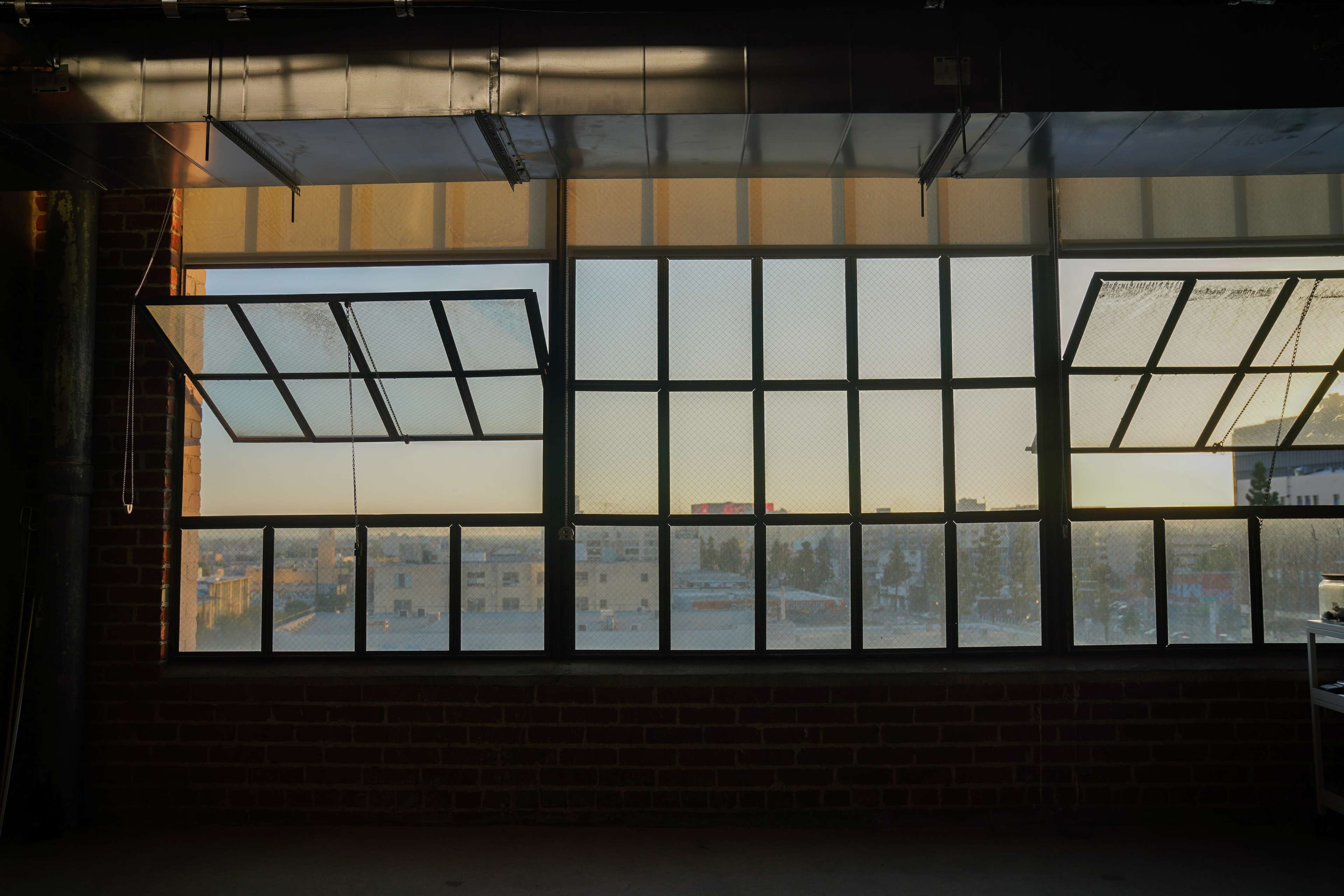 The image shows a large industrial window with two panes open, revealing a view of an urban landscape at sunset.