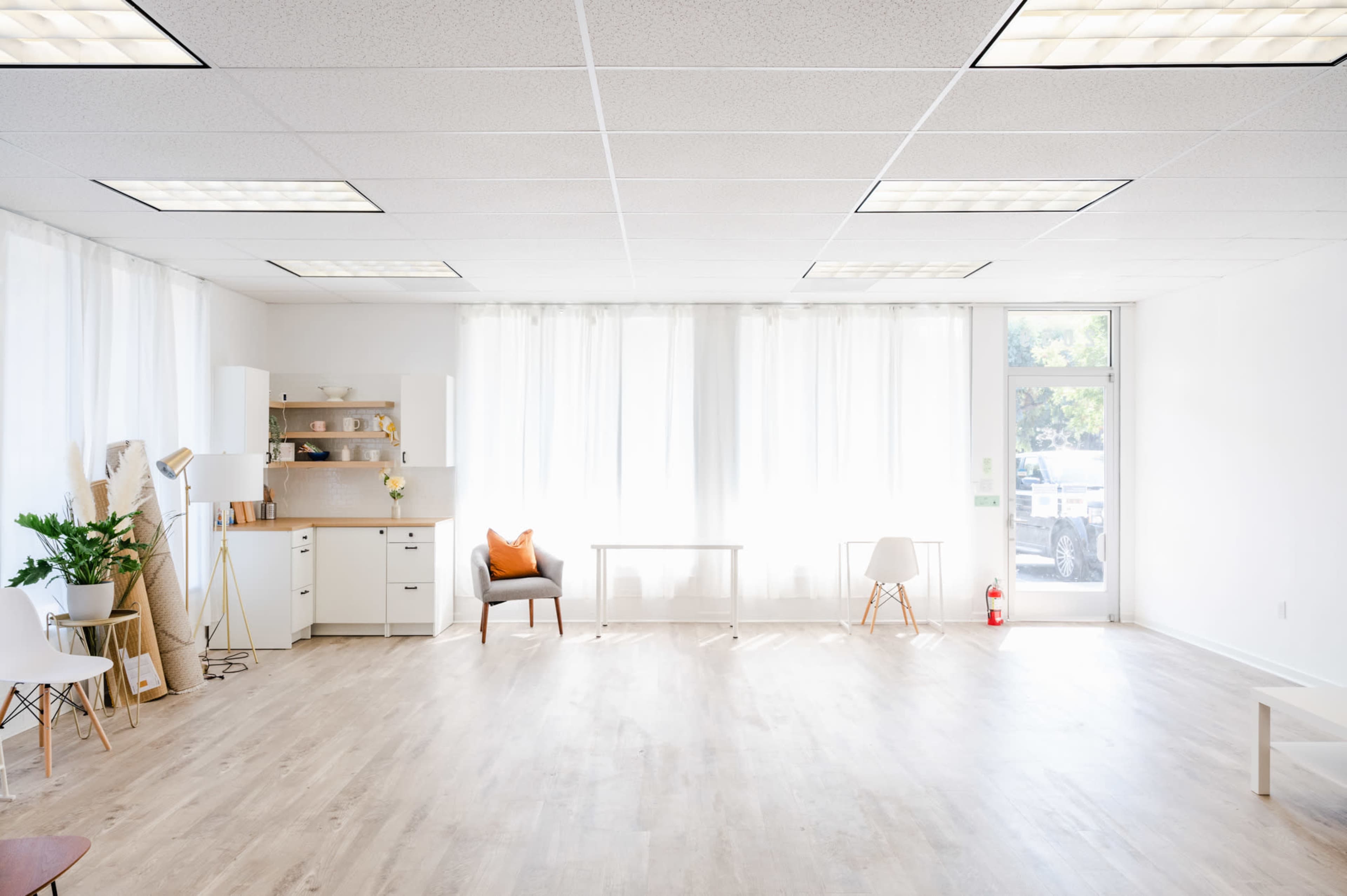 The image shows a bright, spacious interior of an office or studio with white walls, wooden flooring, large windows, and minimal furniture including a table and a chair.