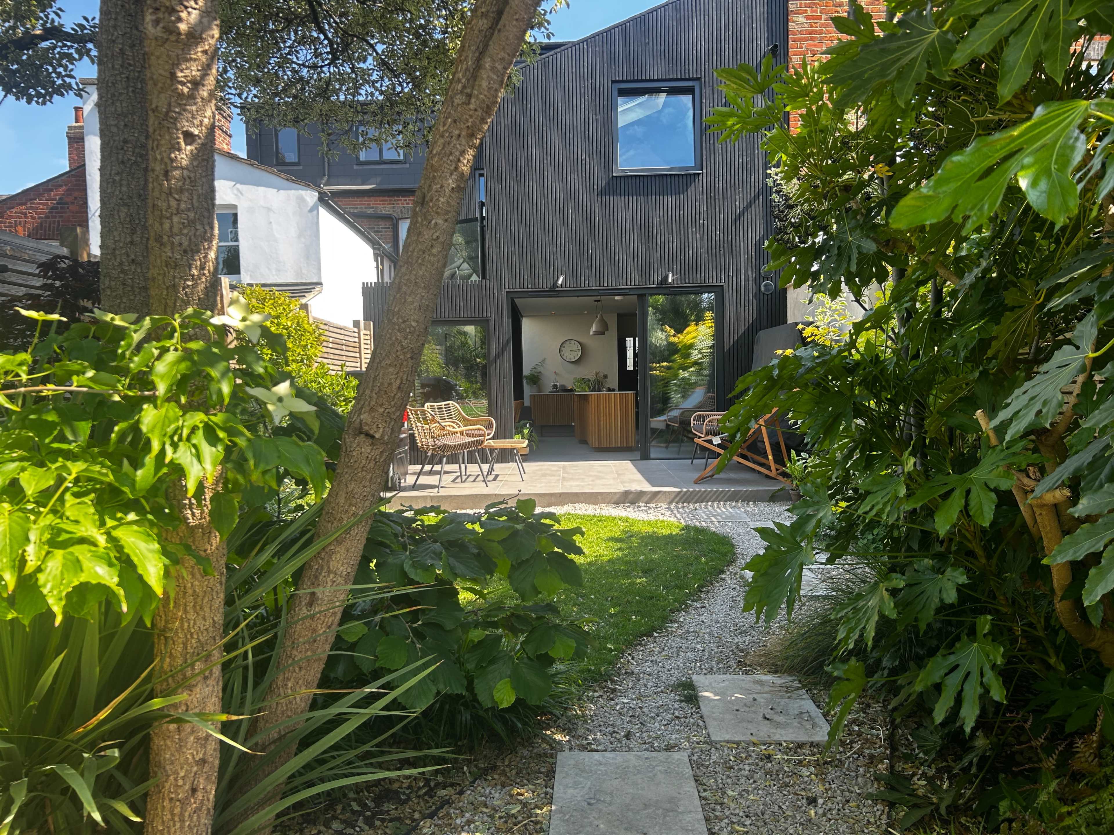 The image shows a modern house with a wooden facade surrounded by lush greenery and a stone pathway leading to the entrance.
