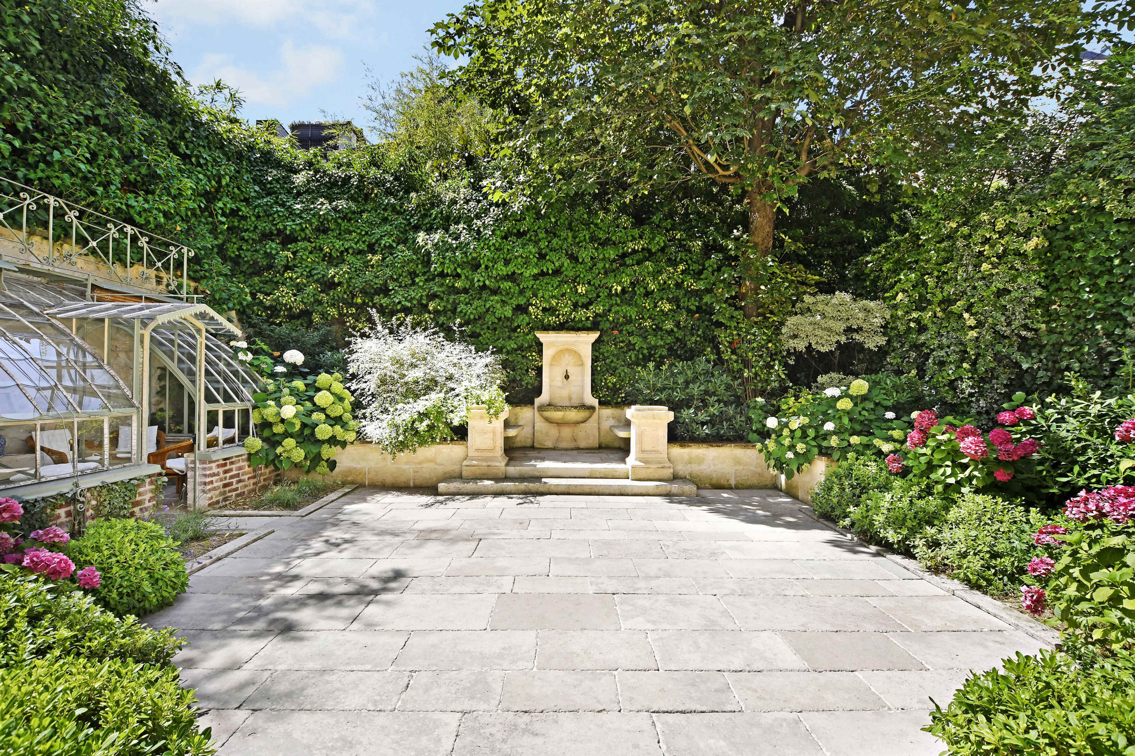 A stone patio surrounded by lush greenery, a fountain, and flowering plants.