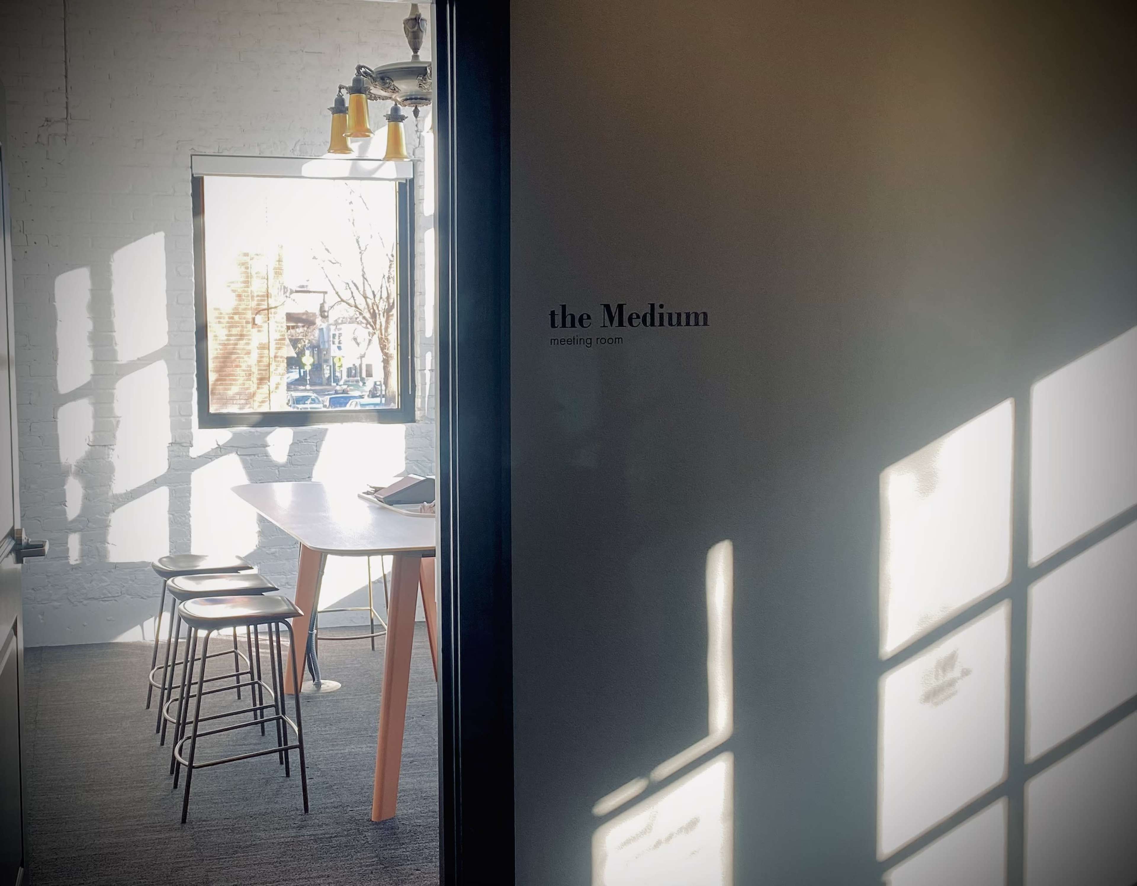 The image shows a modern meeting room labeled "the Medium," featuring a table with chairs and sunlight casting shadows on the wall.
