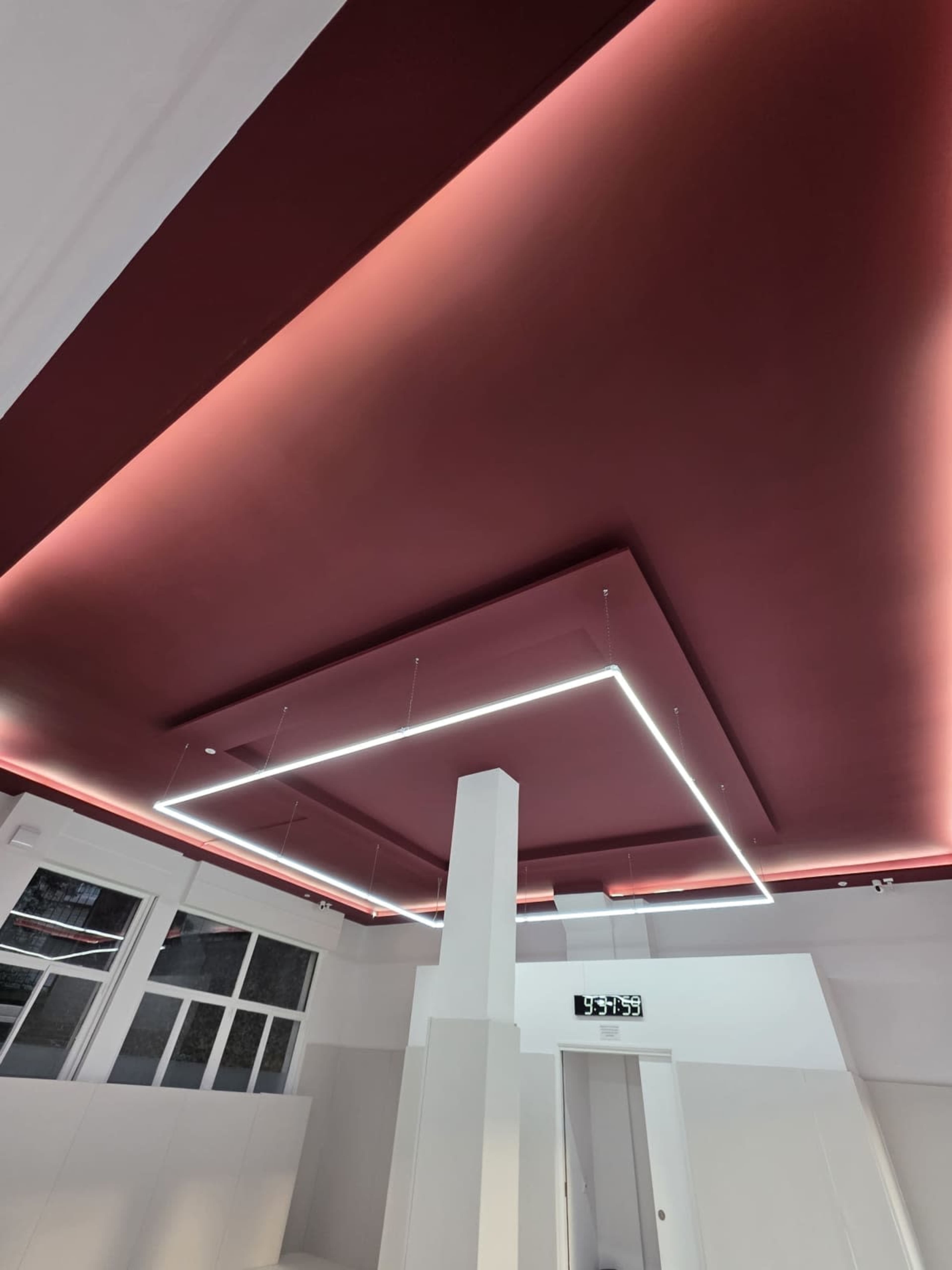 The image shows a modern interior with a maroon ceiling illuminated by geometric LED lighting, and a white column in the center of the space.