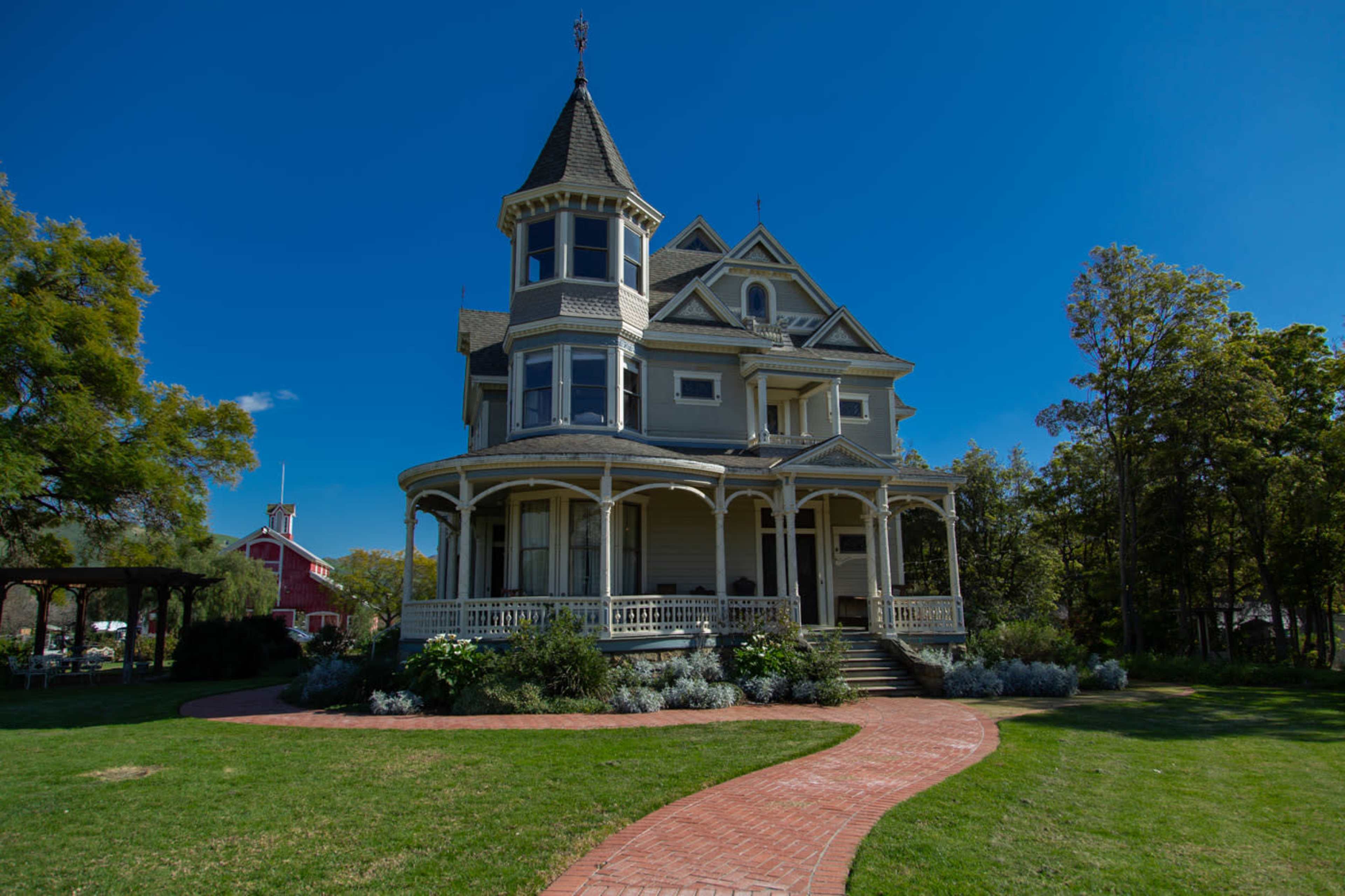 A Victorian-style house with a turret and wraparound porch is situated on a grassy lawn, bordered by a winding brick pathway.