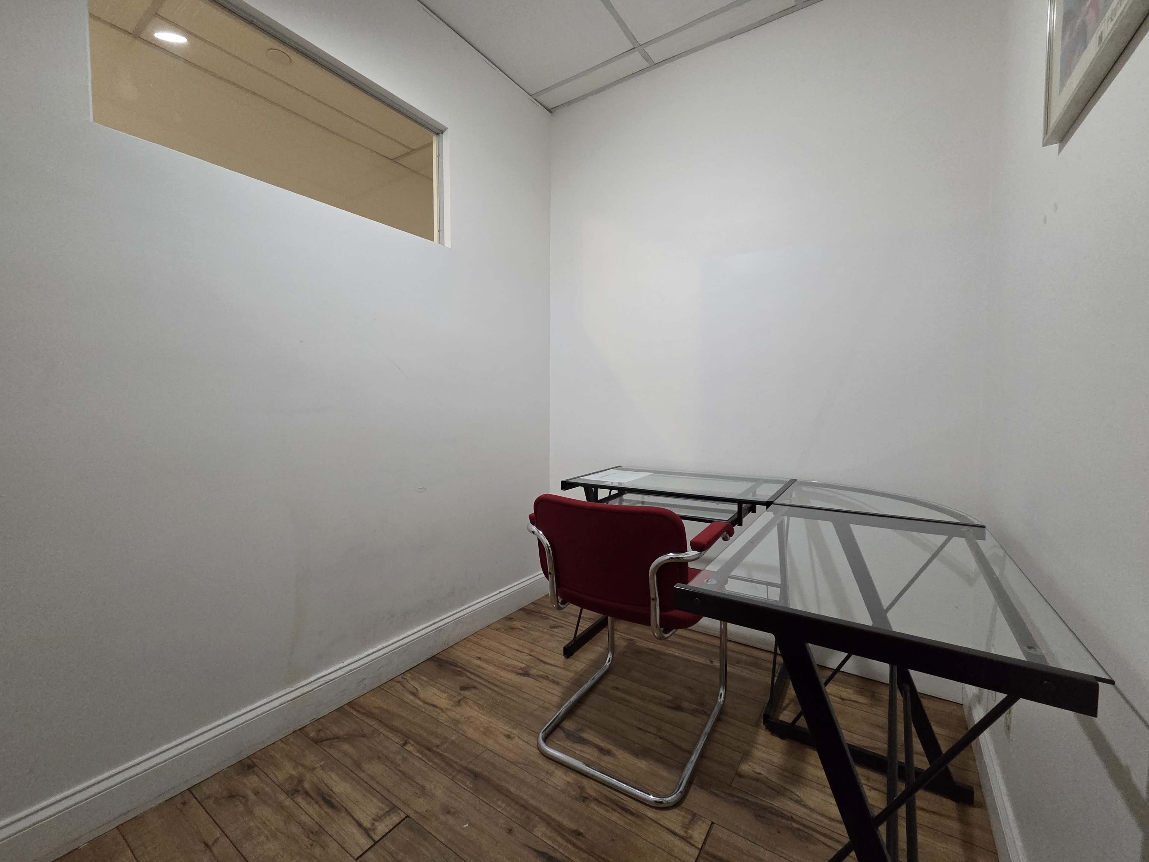 The image shows a small, empty room with two glass-top tables and a red chair against a white wall.