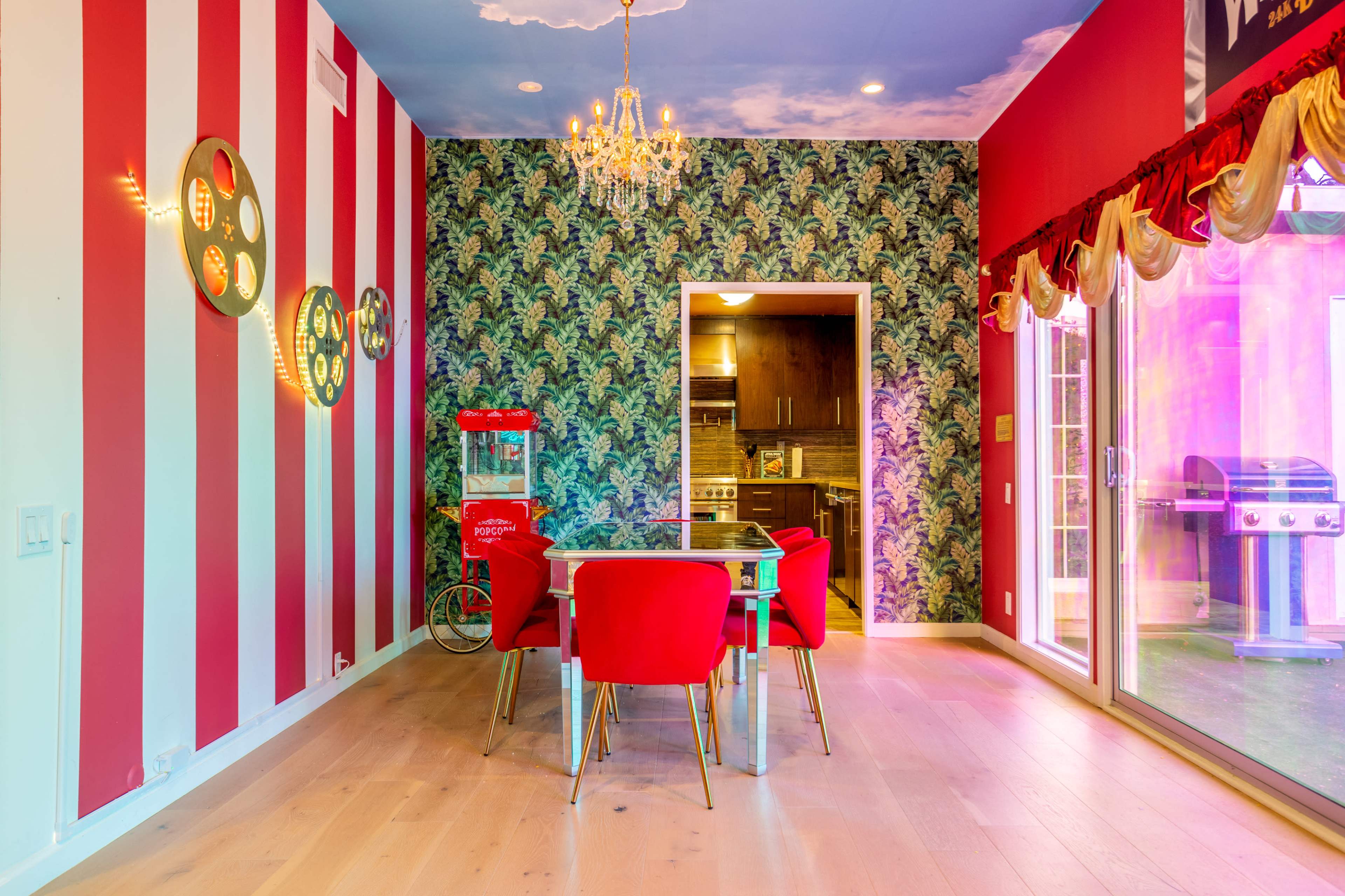The room features red and white striped walls, a tropical patterned wallpaper, a chandelier, and a dining table surrounded by four red chairs.