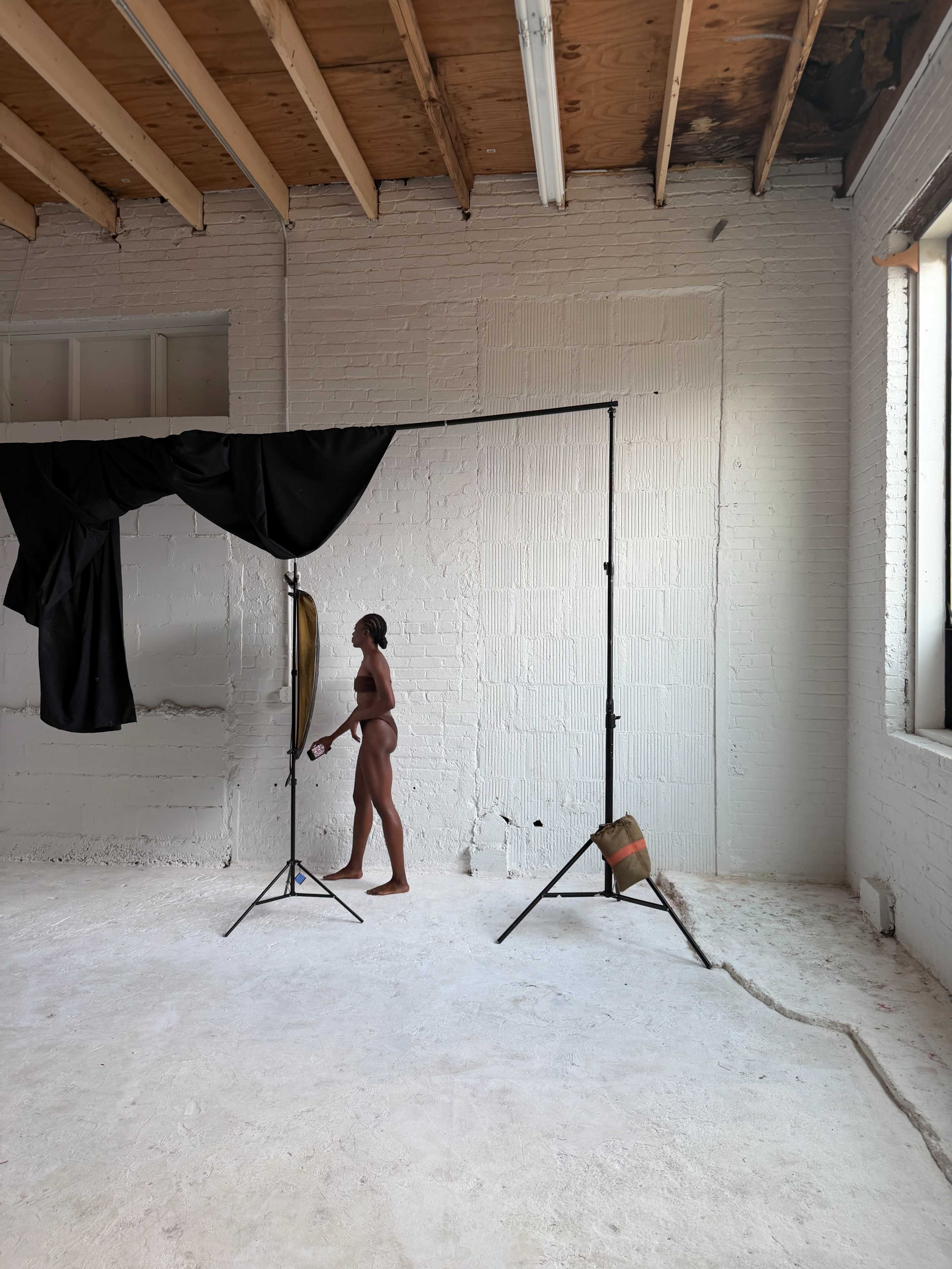 A figure stands alone in a sparse, unfinished studio space with white walls and wooden beams above.