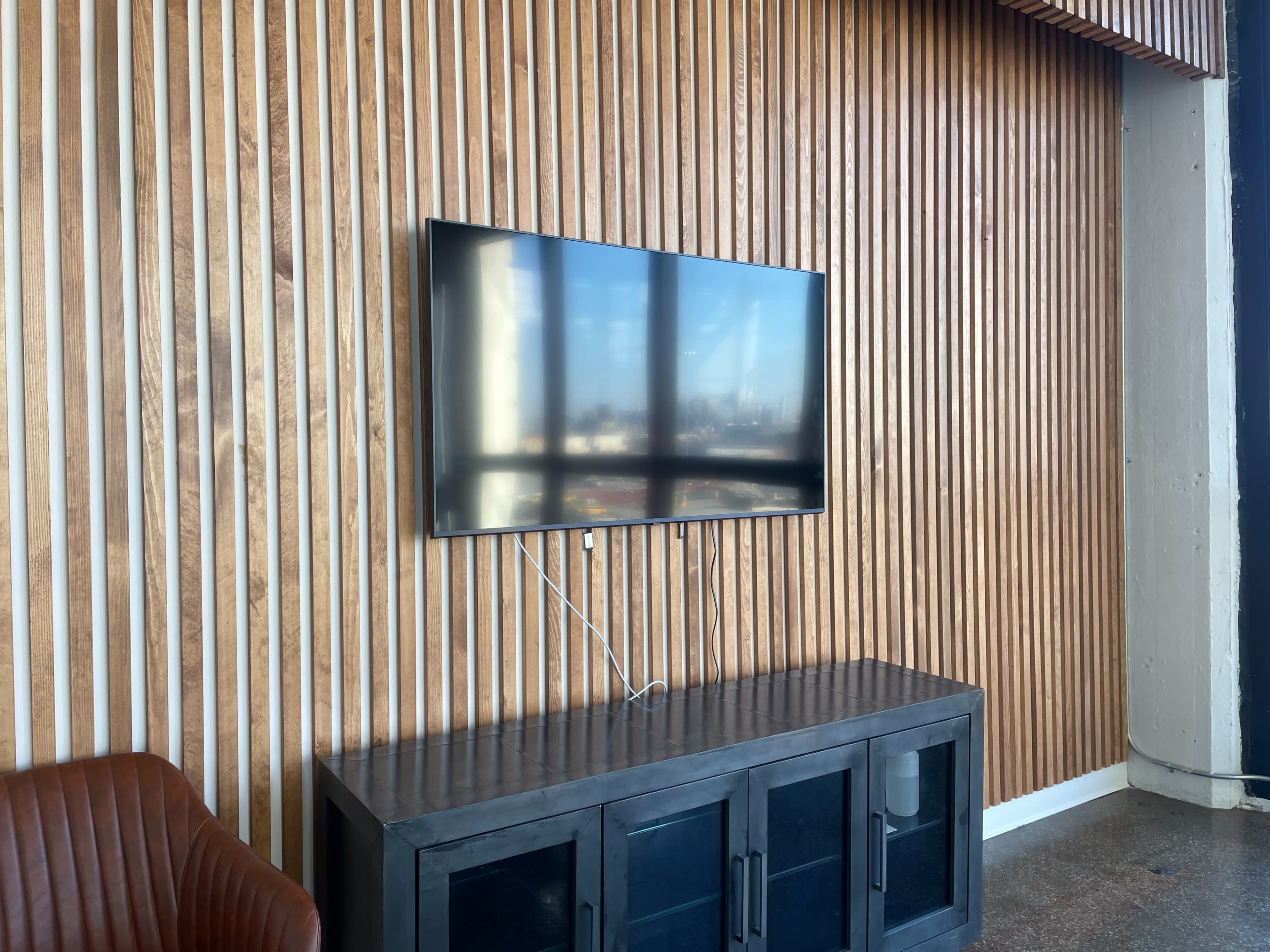 A flat-screen television is mounted on a wooden slat wall above a dark sideboard in a modern interior space.