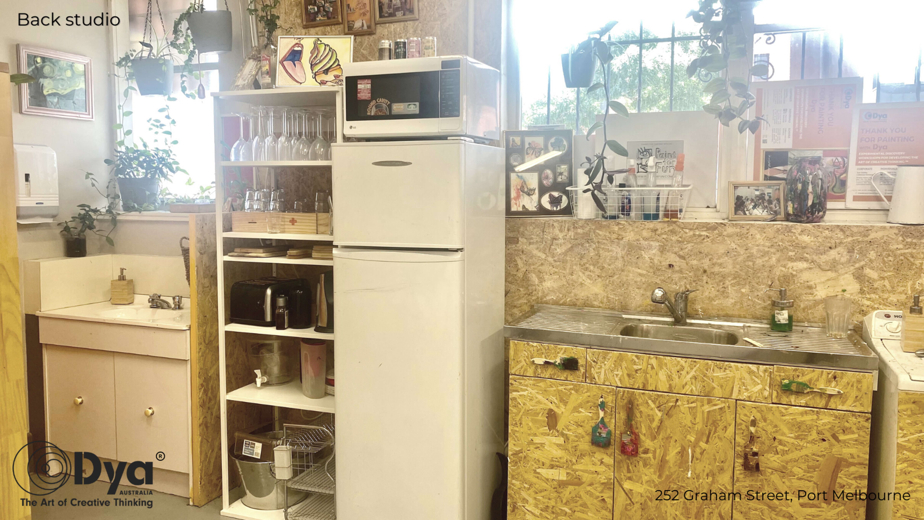 A cluttered kitchen area featuring a white refrigerator, a sink, shelves with glasses and utensils, and a countertop with various appliances in a studio environment.