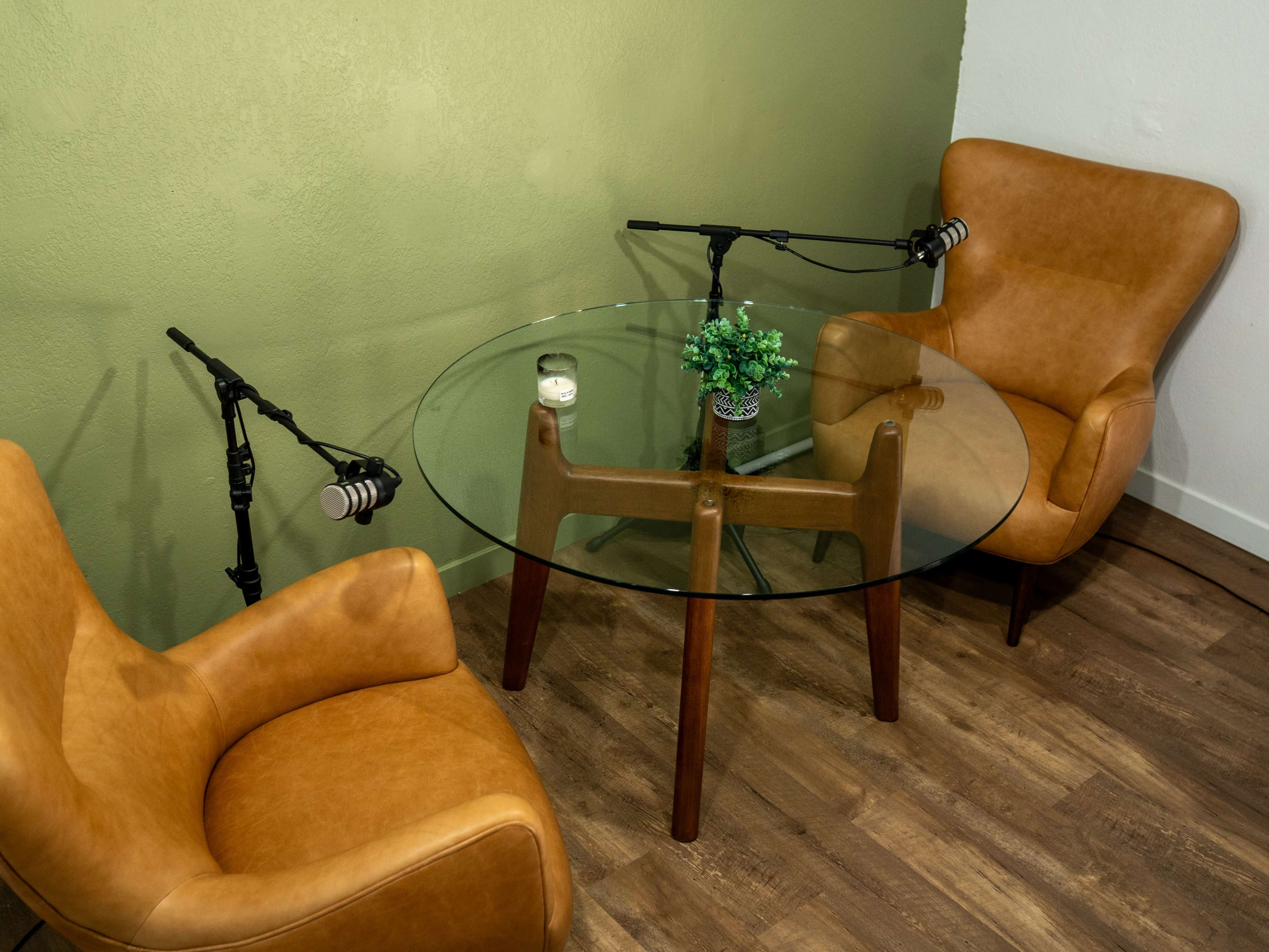 The image shows a small podcast recording setup featuring two brown chairs, a round glass table, and two microphones mounted on stands against a green wall.