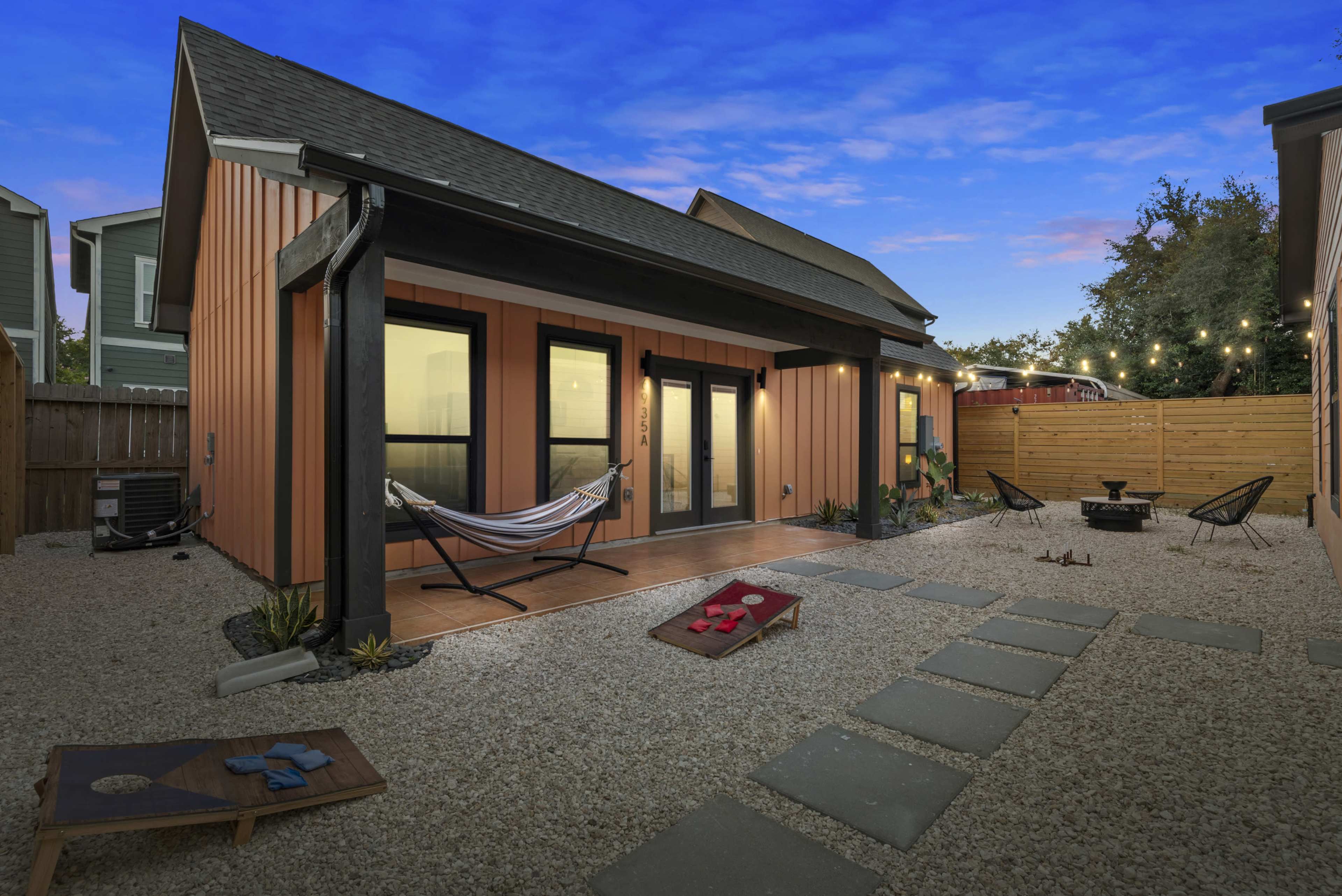 The image shows a modern outdoor area with a gravel patio featuring a hammock, a fire pit, and a pathway lined with stone slabs leading to a house with orange and black siding.