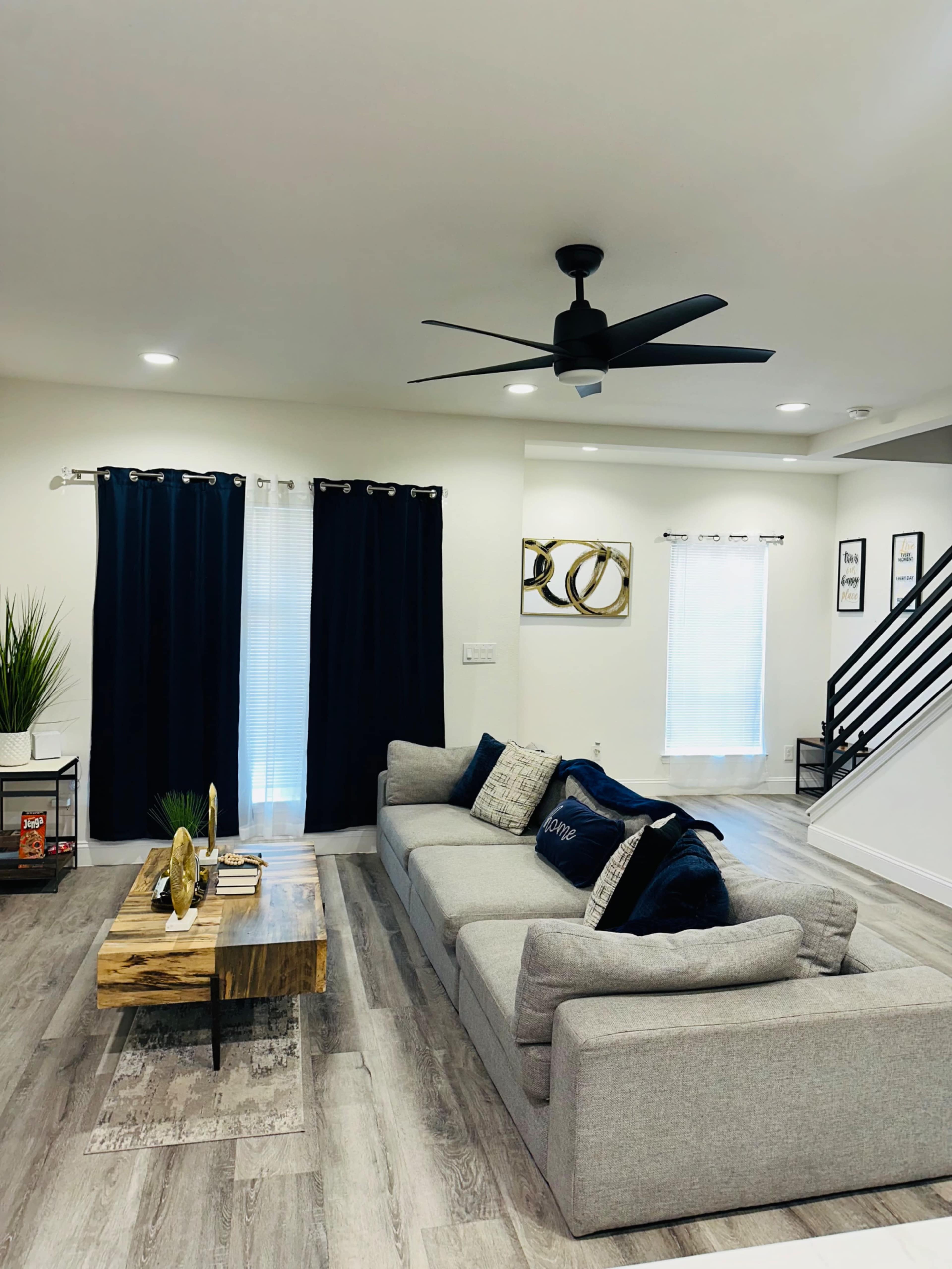 A modern living room features a gray sectional sofa, a wooden coffee table, dark curtains, and a ceiling fan, with stairs and wall art in the background.