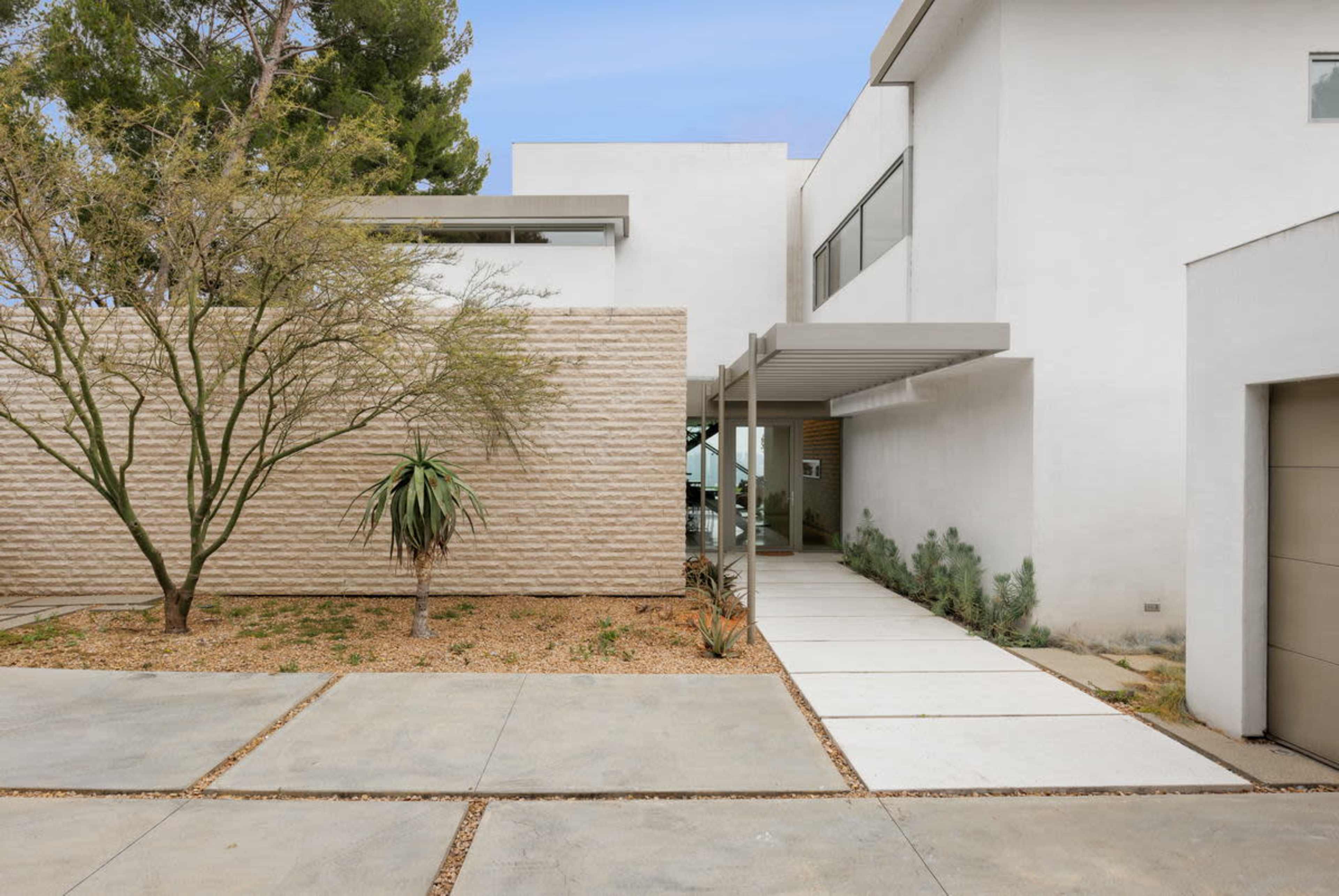A modern house with a light-colored exterior features a landscaped front yard and a concrete pathway leading to the entrance.