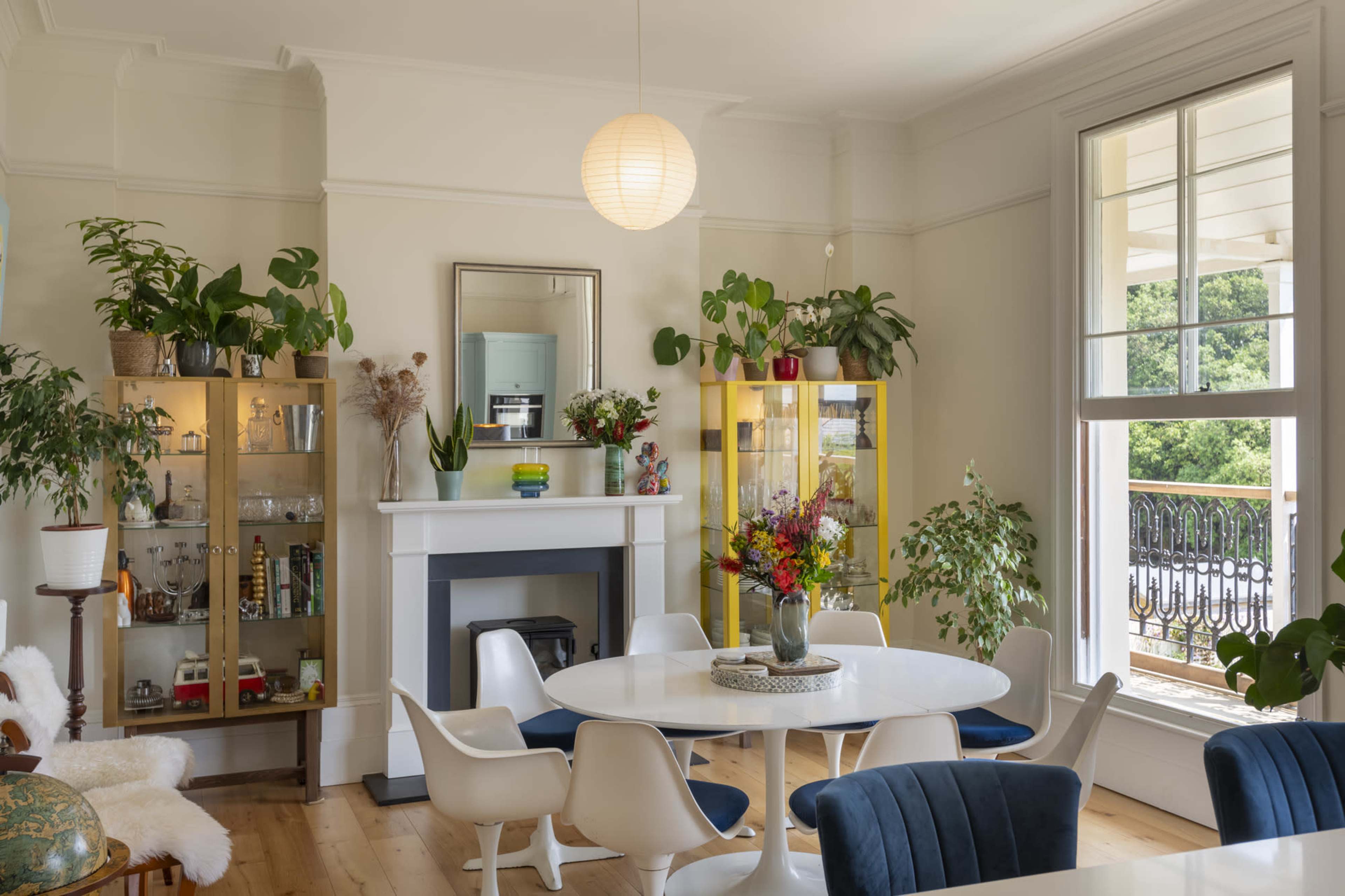 The image shows a modern dining area with a round table, blue chairs, and two display cabinets filled with various items, all accented by large windows and indoor plants.