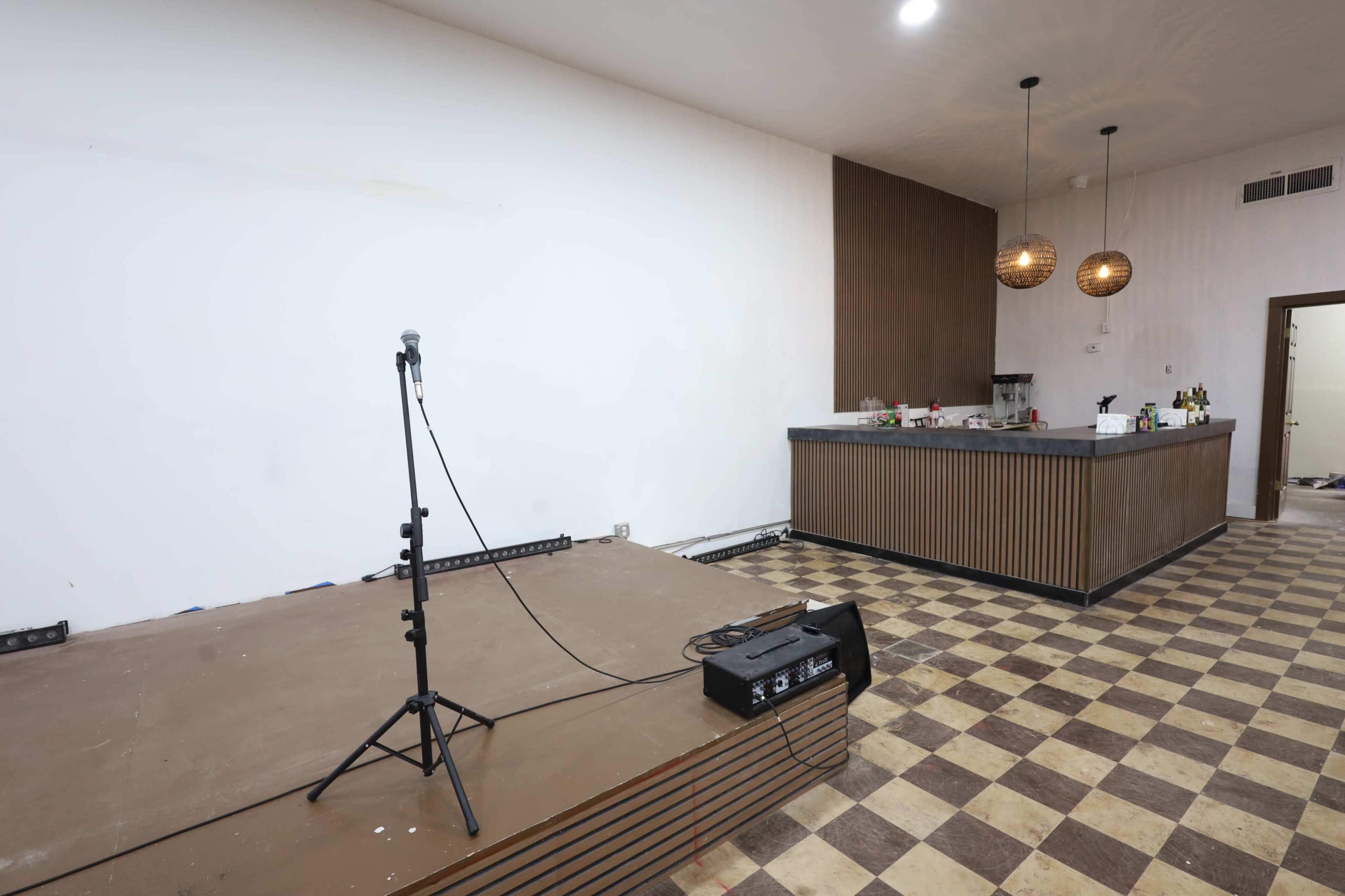 The image shows a partially furnished room with a microphone stand on a small stage and a bar area that includes a counter with several bottles.