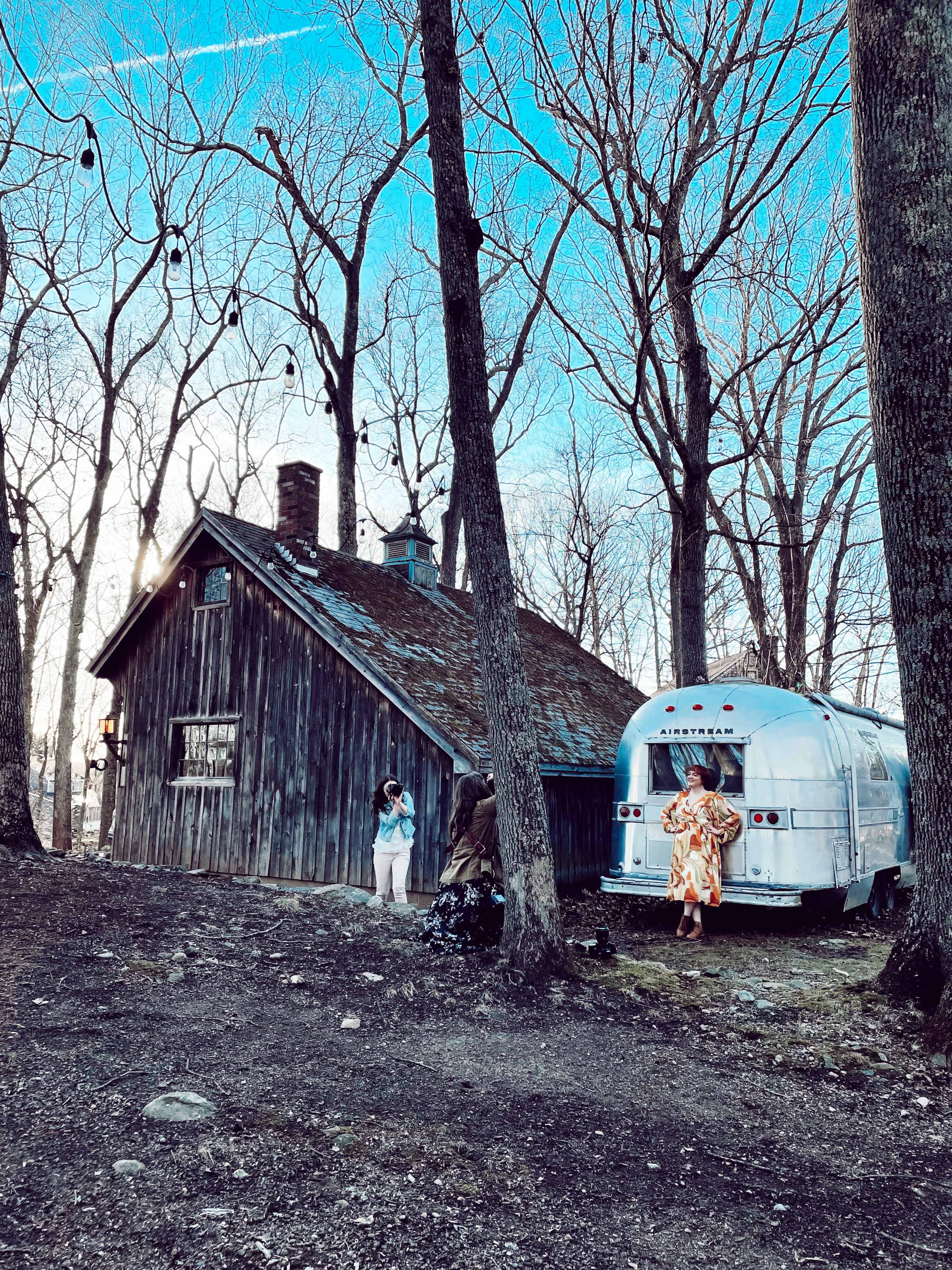 A silver Airstream trailer is parked next to a rustic wooden cabin surrounded by bare trees.