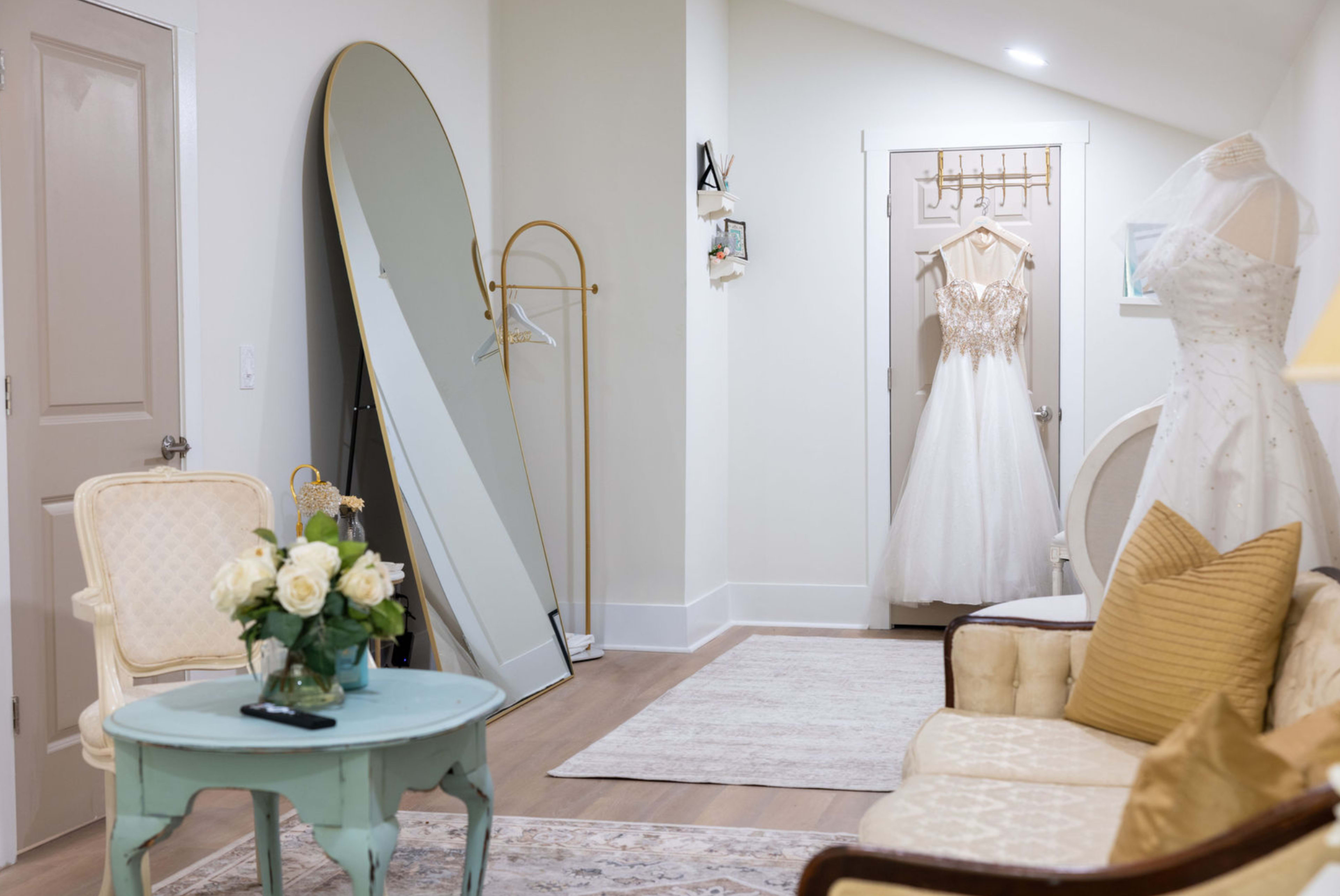 A well-lit dressing room features a large mirror, a hanging wedding dress, and a seating area with a small table and floral centerpiece.