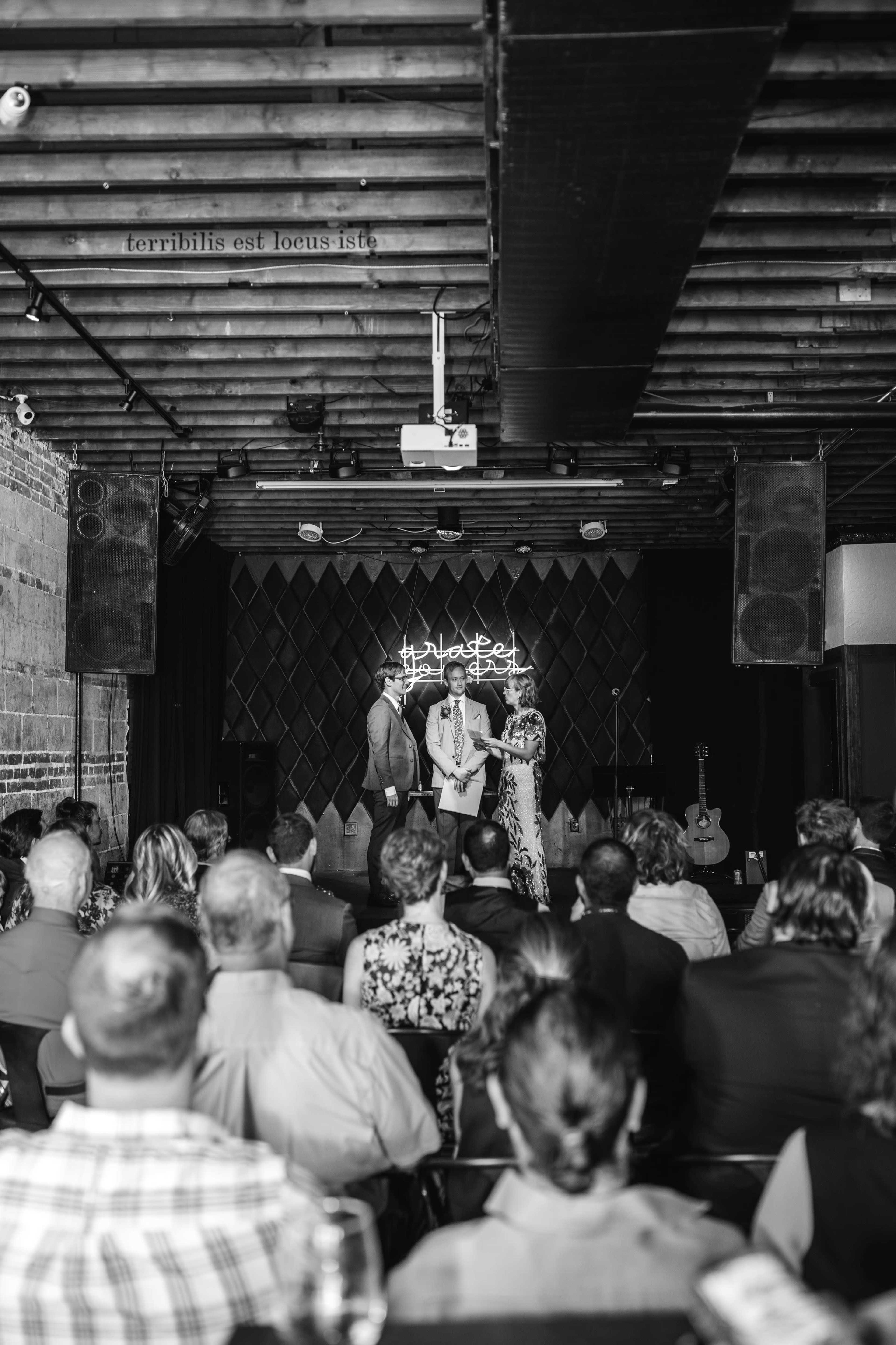 A group of people sits in an audience while three individuals stand on a stage in a dimly lit venue.
