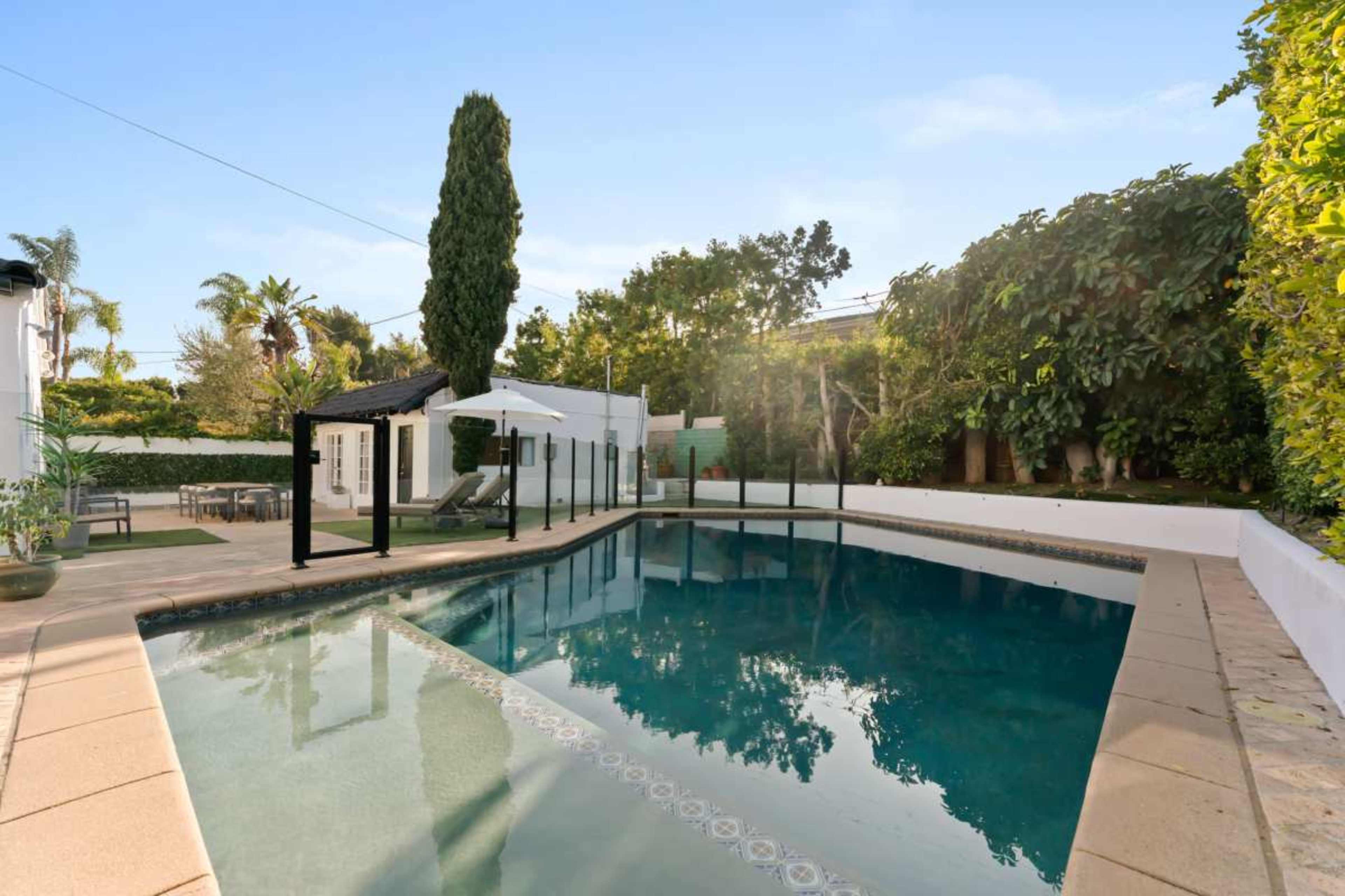 The image shows a backyard pool area enclosed by a fence, surrounded by trees and greenery, with a sunlit sky above.