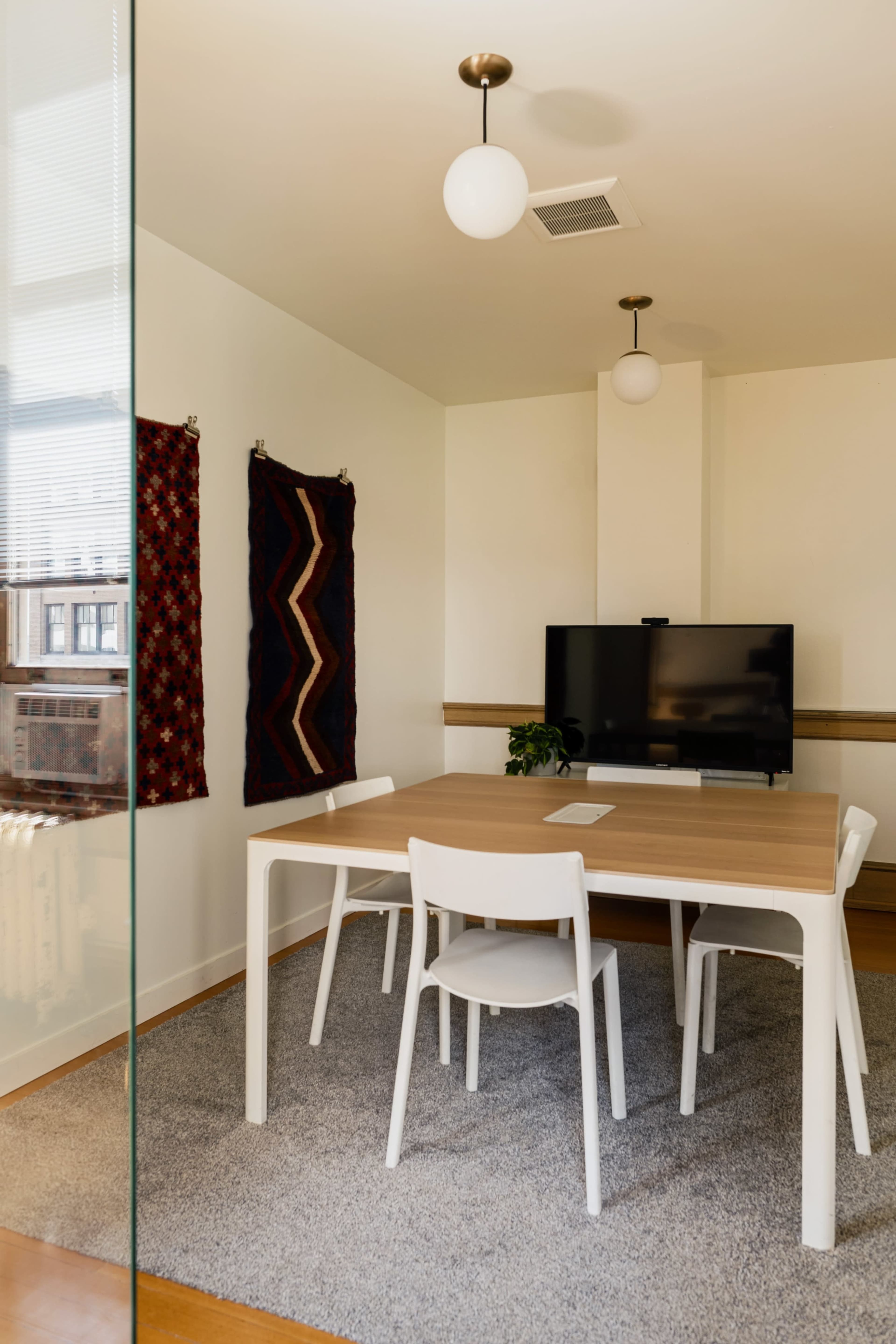 A modern office meeting room features a wooden table surrounded by white chairs, a television on the wall, and two decorative wall hangings.