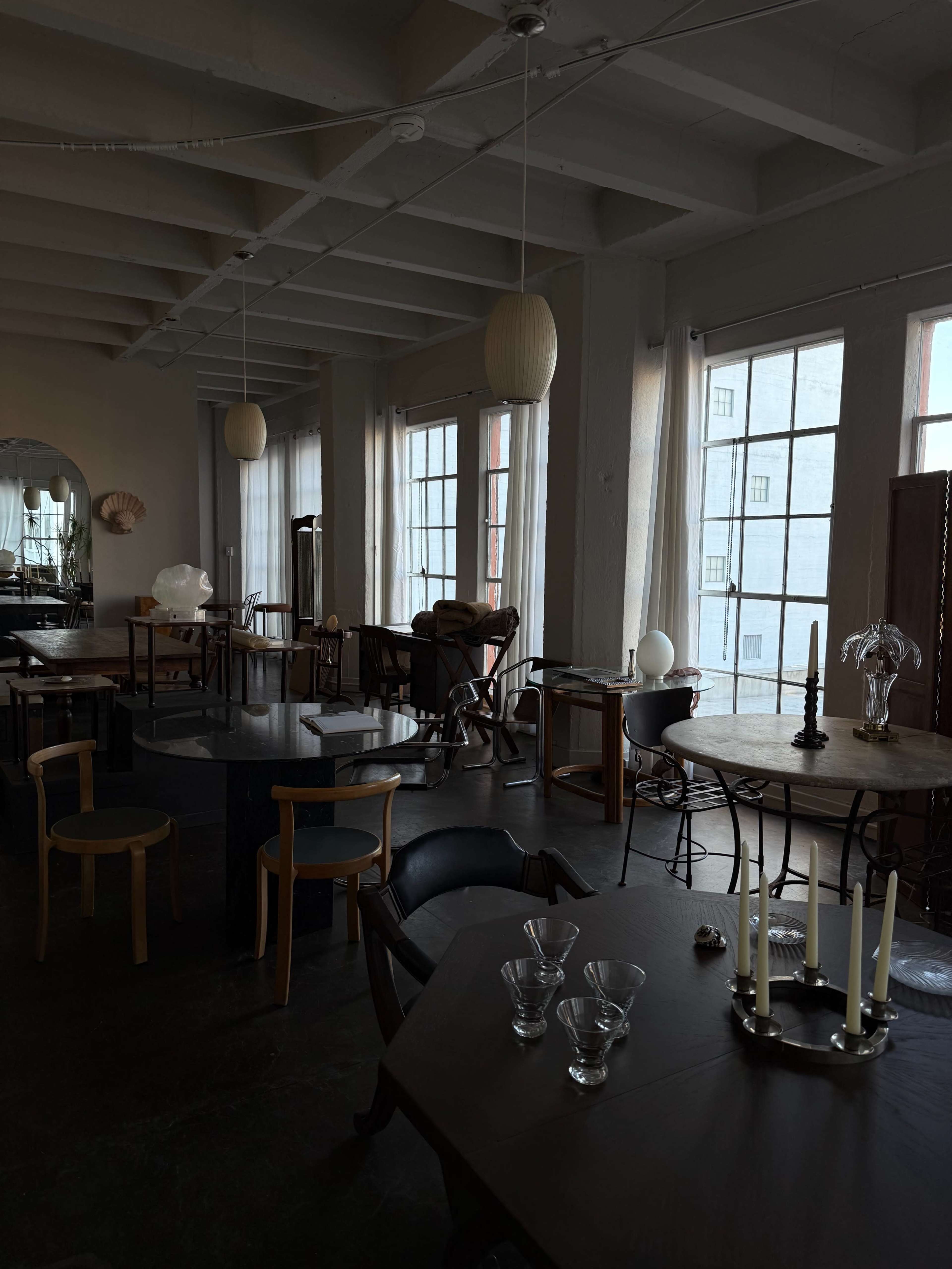 The image shows a spacious interior featuring various tables and chairs, with large windows allowing natural light to fill the room.