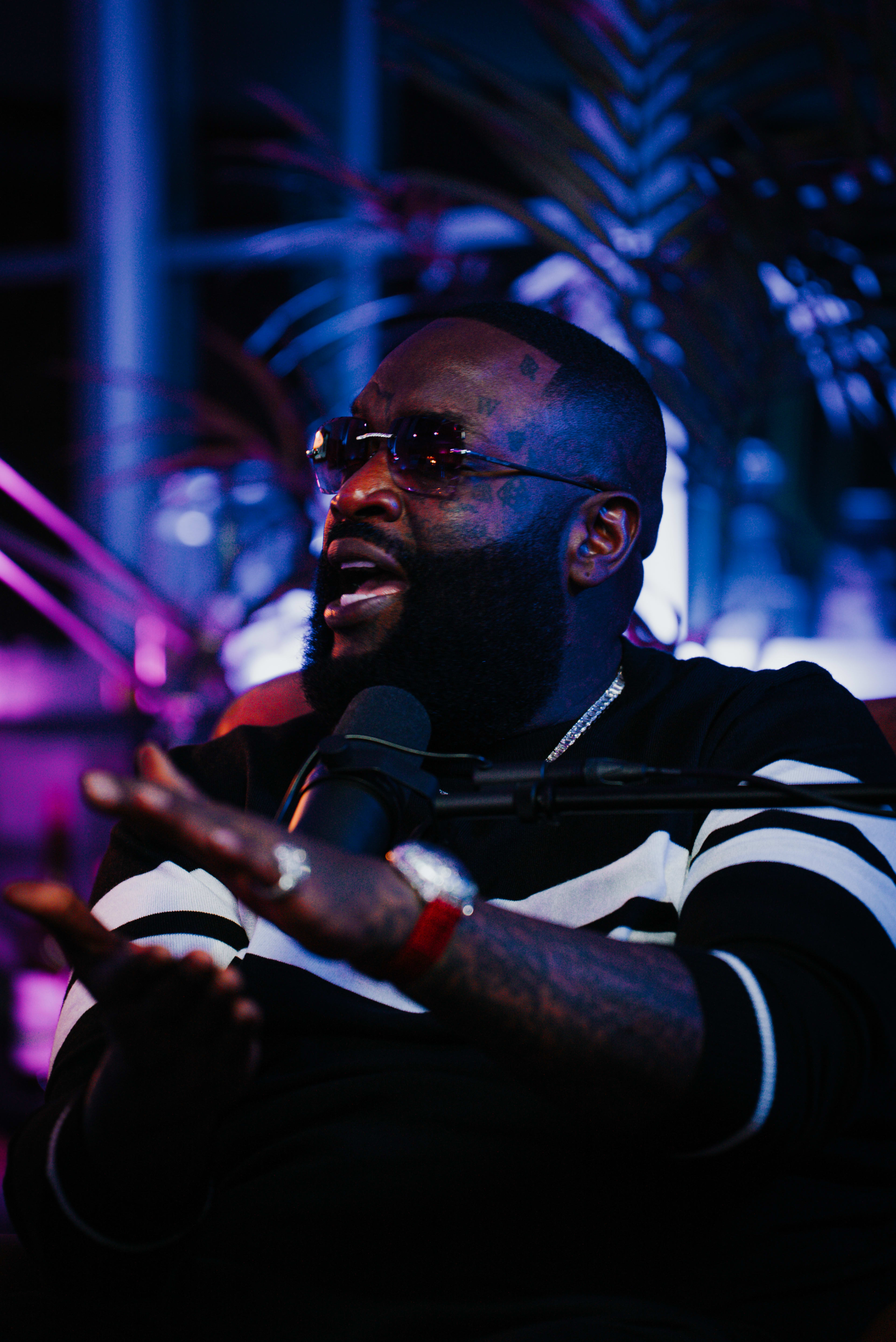 A man wearing sunglasses and a black sweater is speaking animatedly into a microphone in a dimly lit room with colorful lighting and plants in the background.