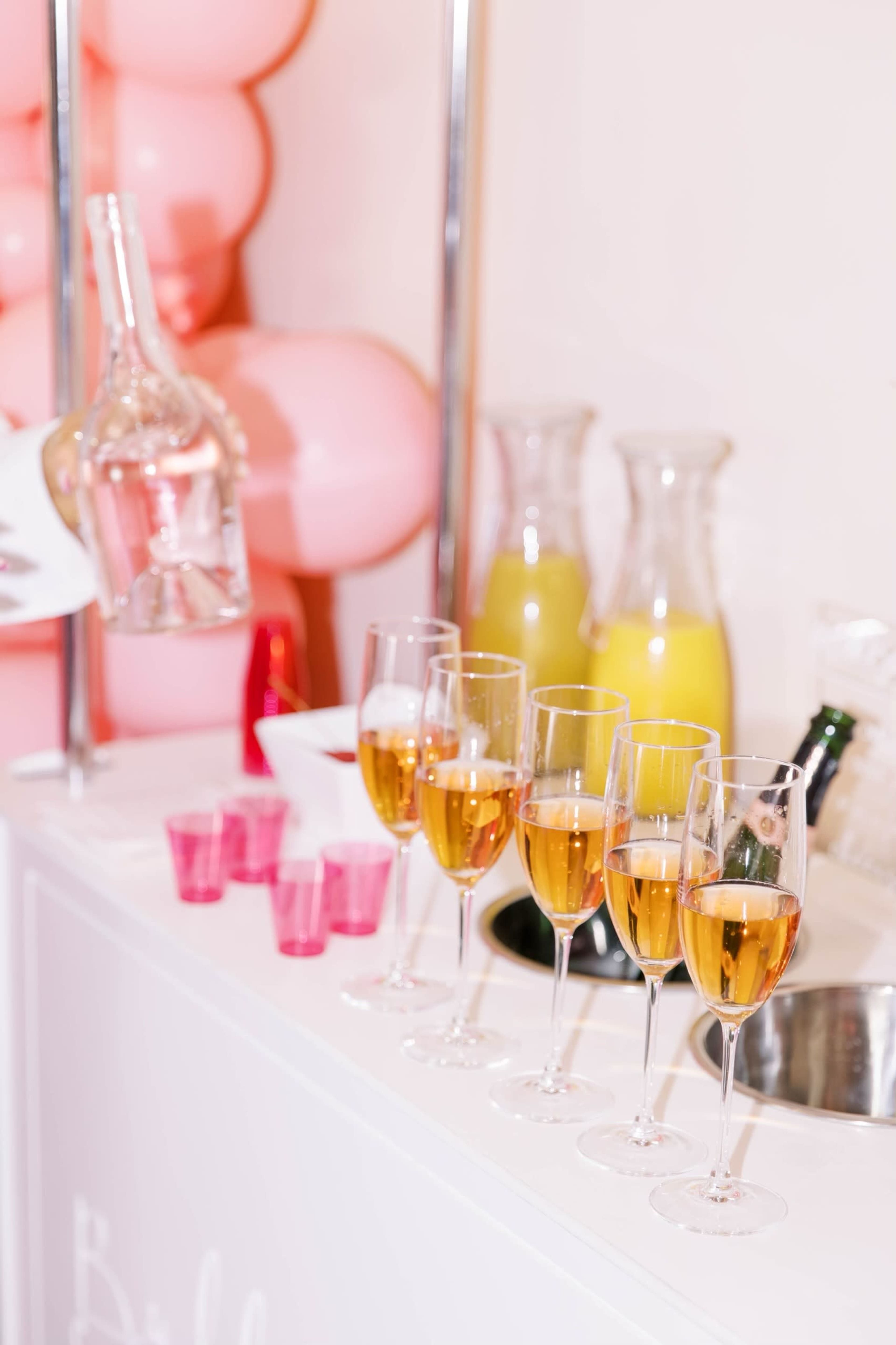 A bar countertop displays several glasses of champagne alongside pitchers of yellow liquid and pink shot glasses.