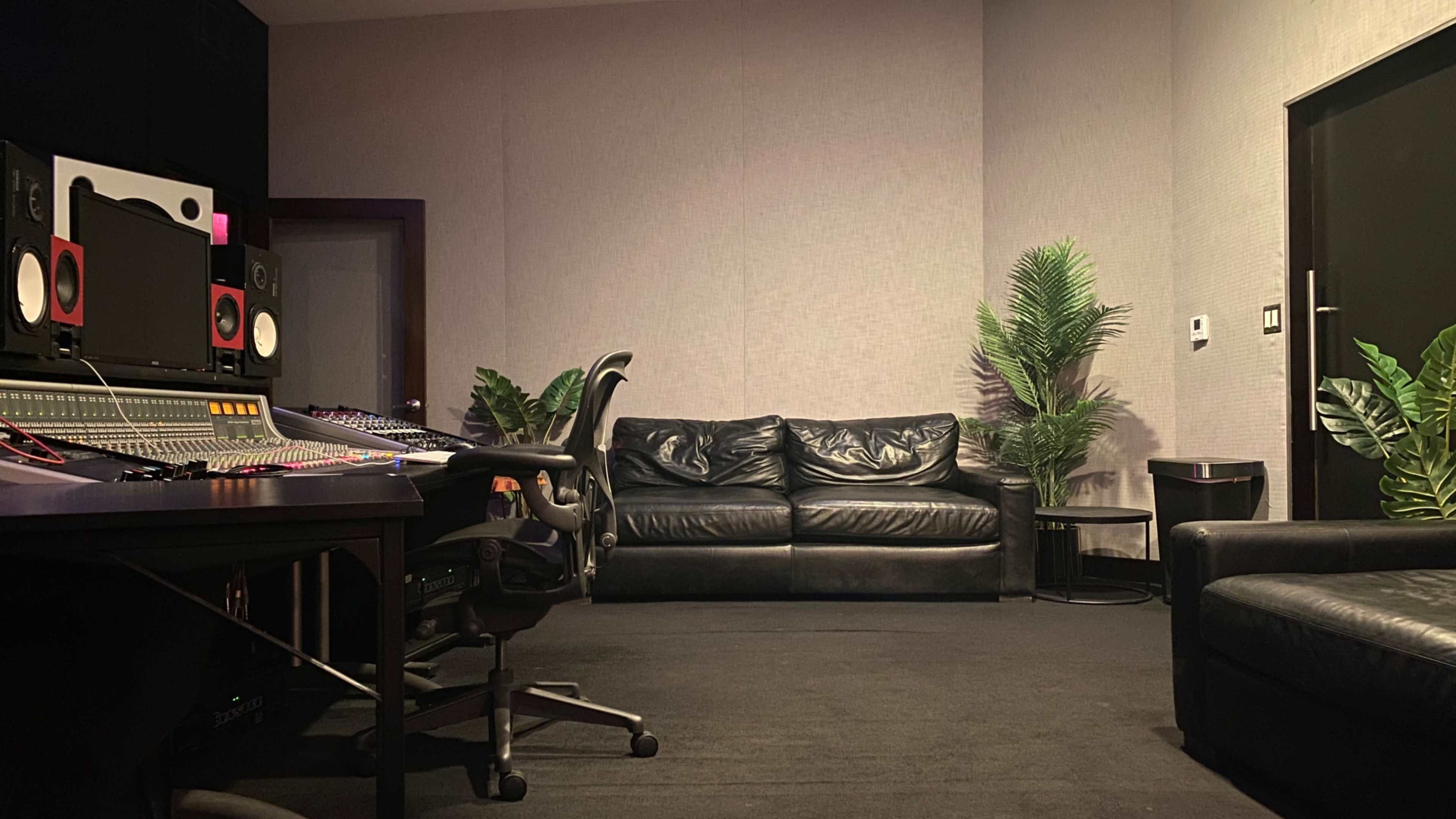 The image shows a recording studio with a large sound mixing console, a black leather sofa, and potted plants against the wall.