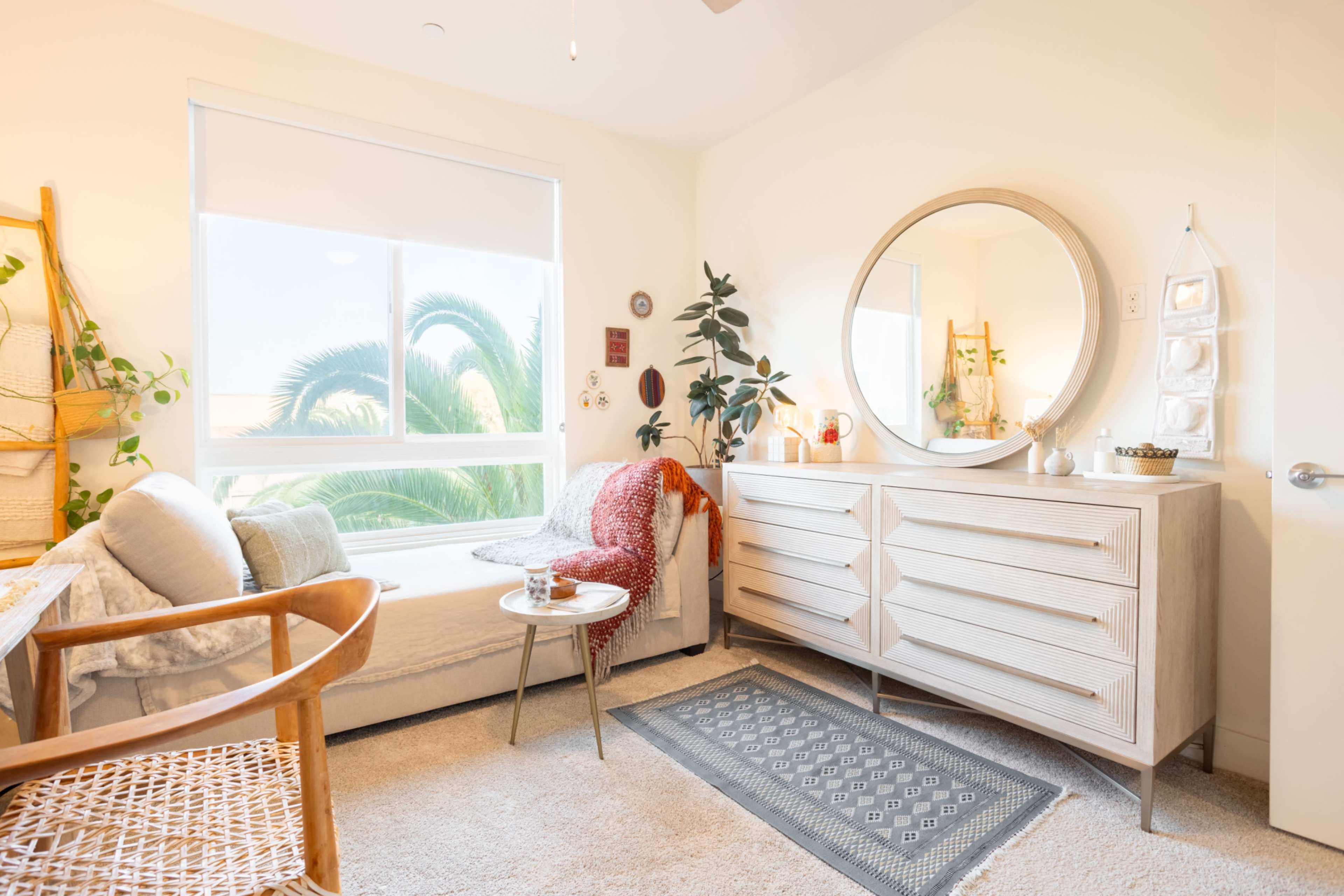 The image shows a bright, minimalist bedroom featuring a beige sofa, a round mirror, and a light wooden dresser alongside a small table and decorative plants.