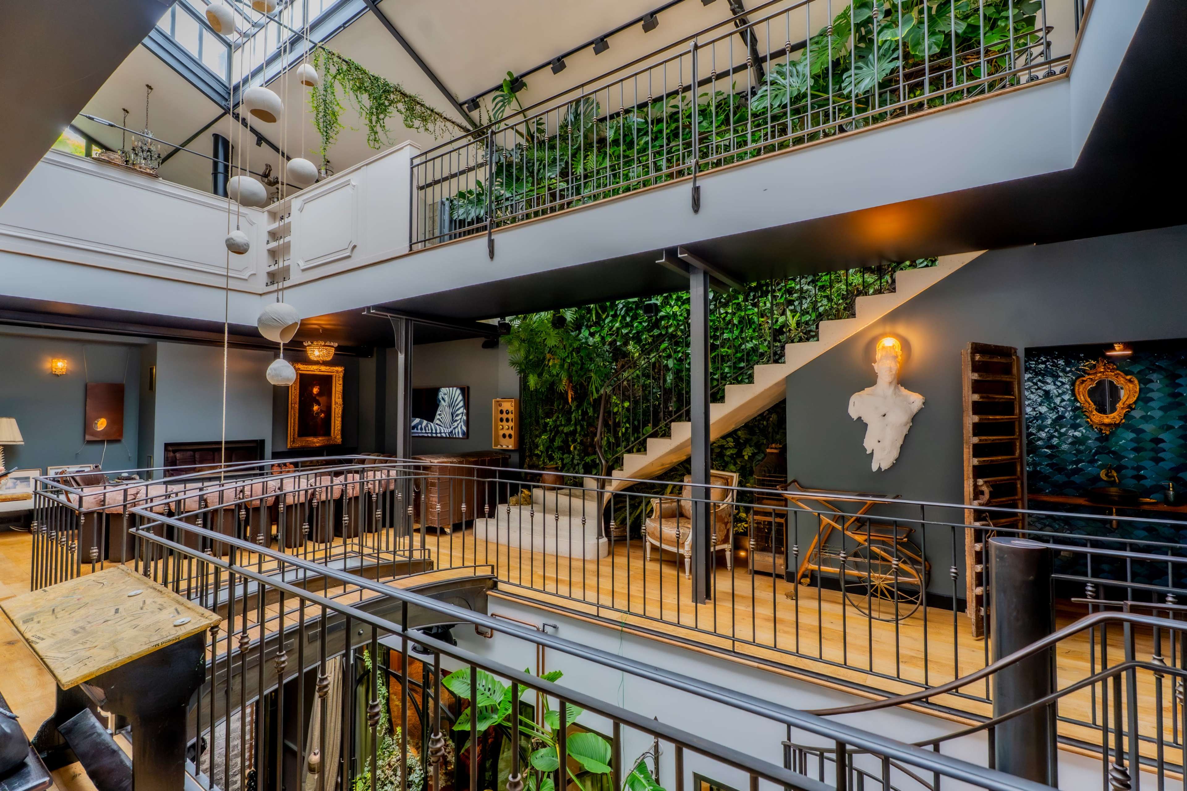 The image depicts an interior space featuring multiple levels connected by a staircase, lush greenery along the walls, and modern lighting fixtures.