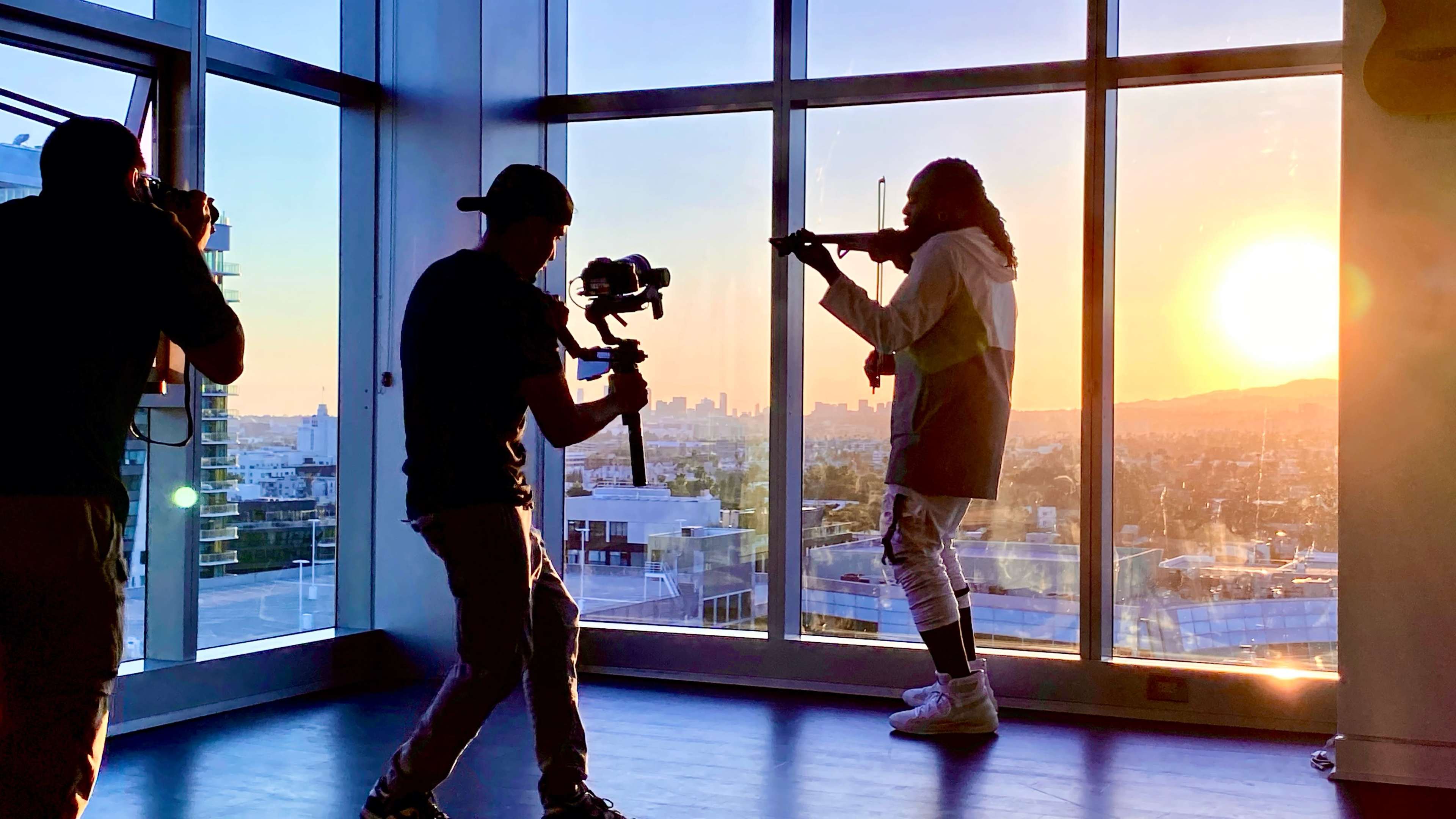 A silhouette of a musician playing the violin is captured against a sunset while a cameraman films the scene.
