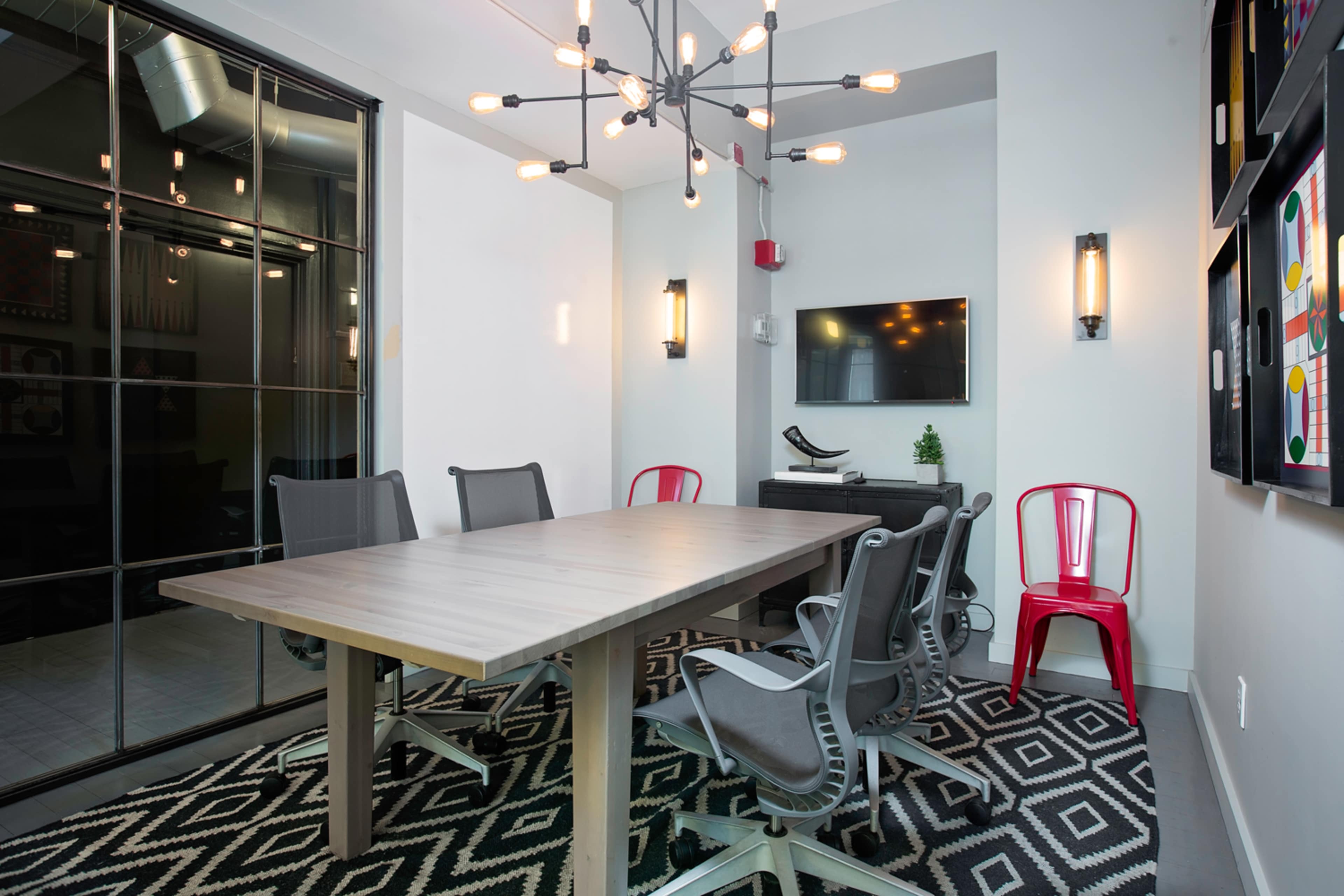 A modern conference room features a long wooden table surrounded by six gray chairs, a red chair, and a chandelier overhead, with a television mounted on the wall and glass-paneled doors.