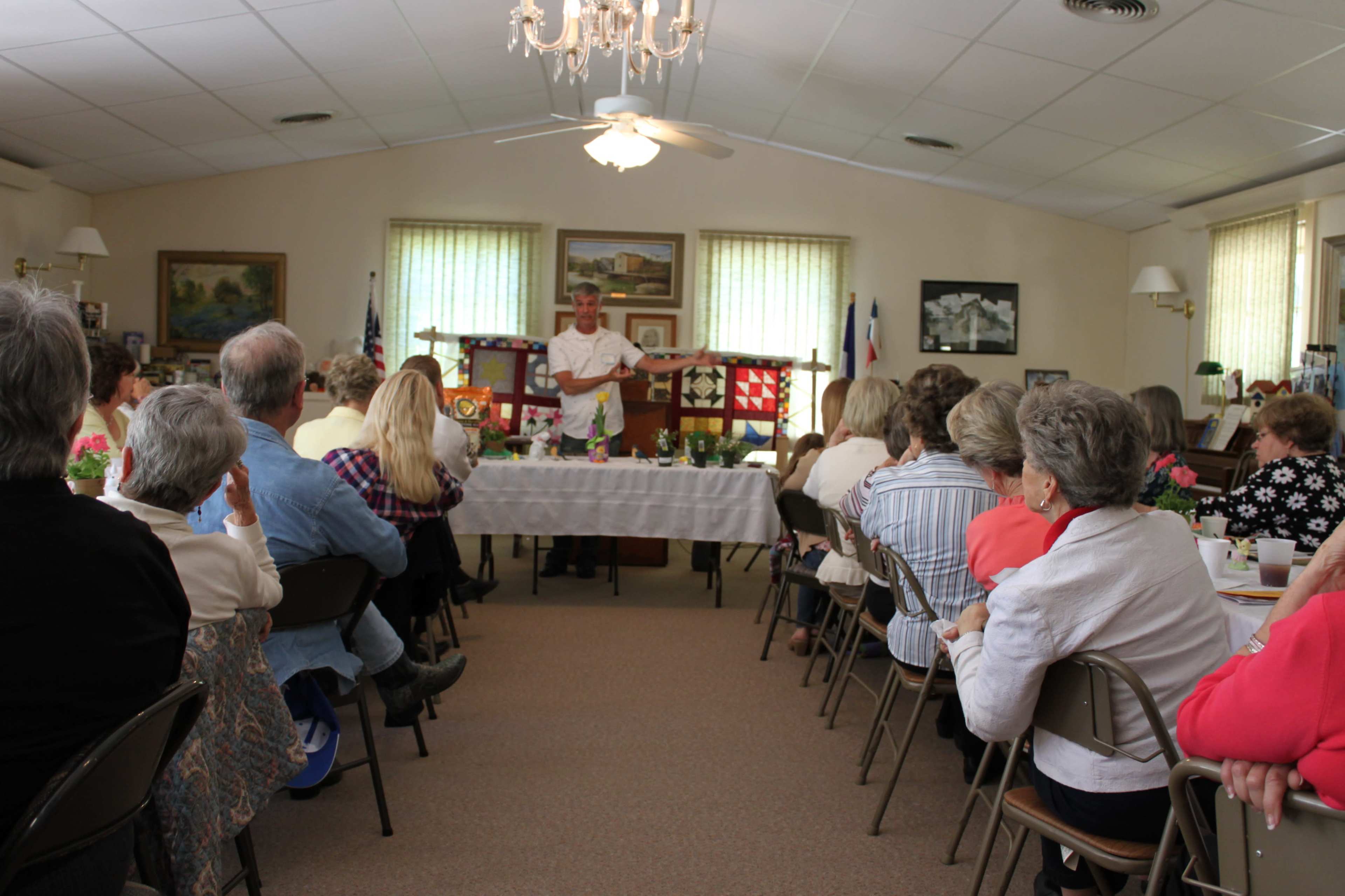 A speaker presents at a gathering in a community hall, with attendees seated at tables arranged in rows.