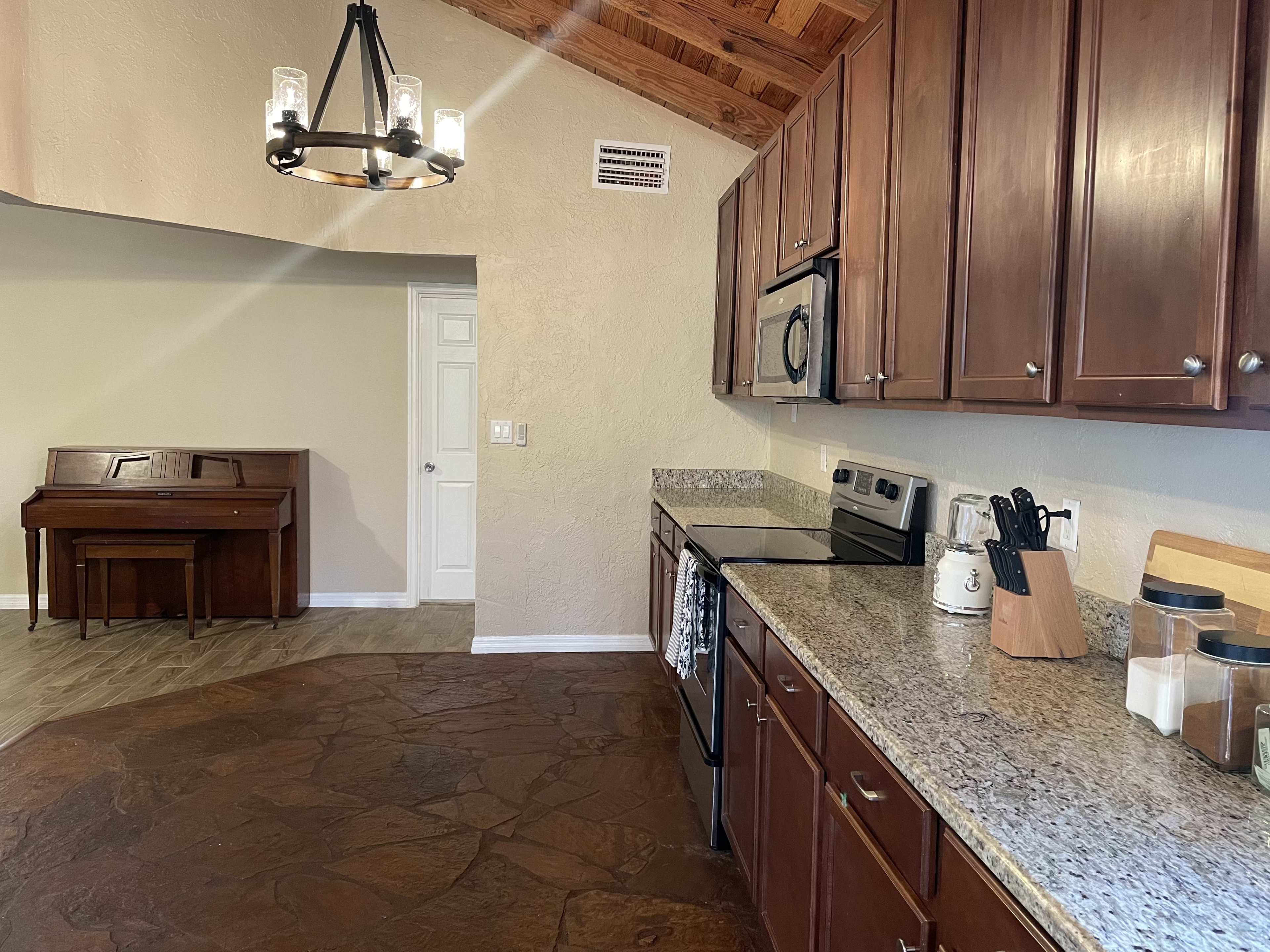 A kitchen with wooden cabinets, granite countertops, and stainless steel appliances, adjacent to a room featuring a piano.