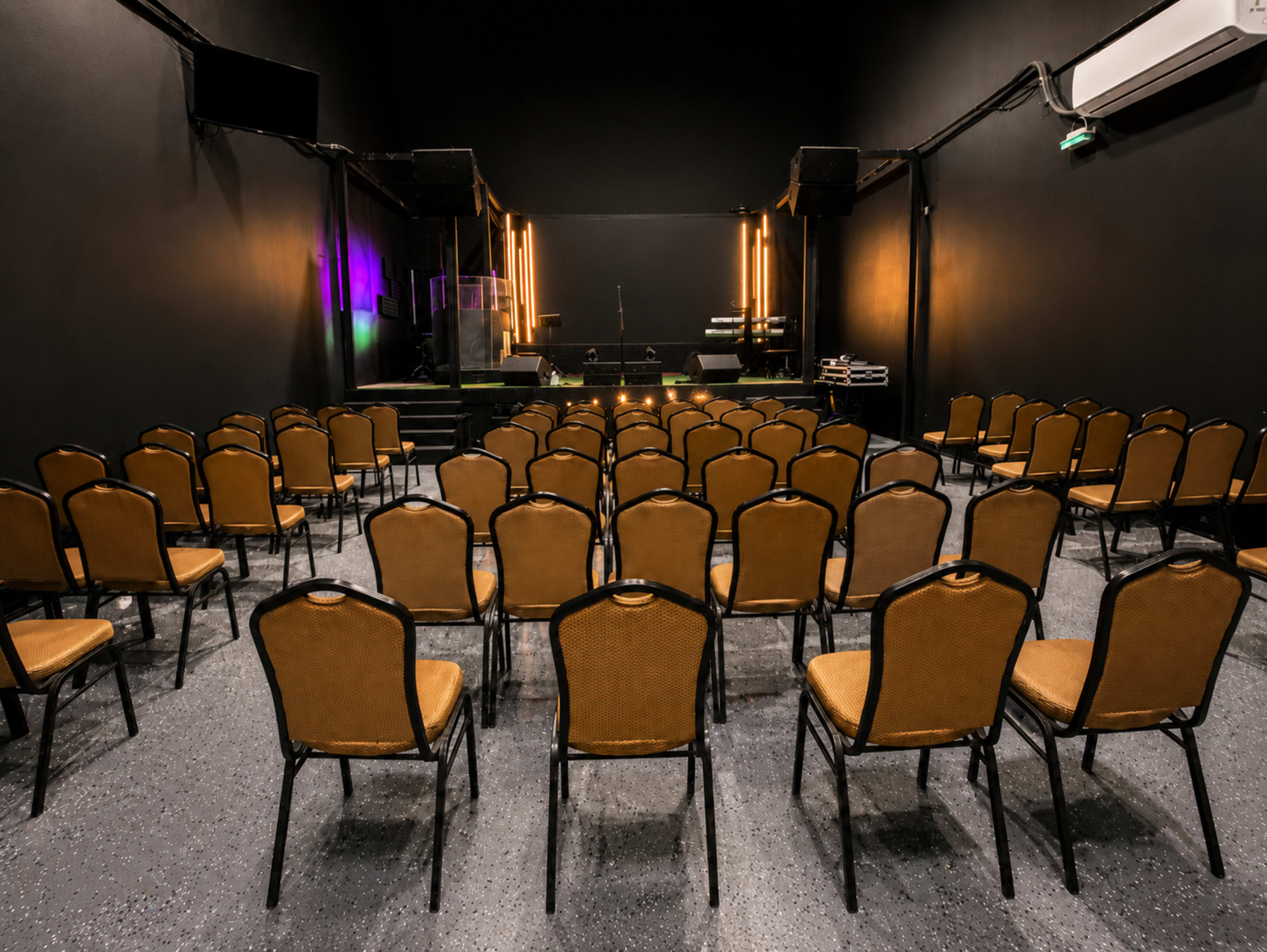 The image shows a concert venue with rows of empty chairs facing a stage illuminated by a few lights.