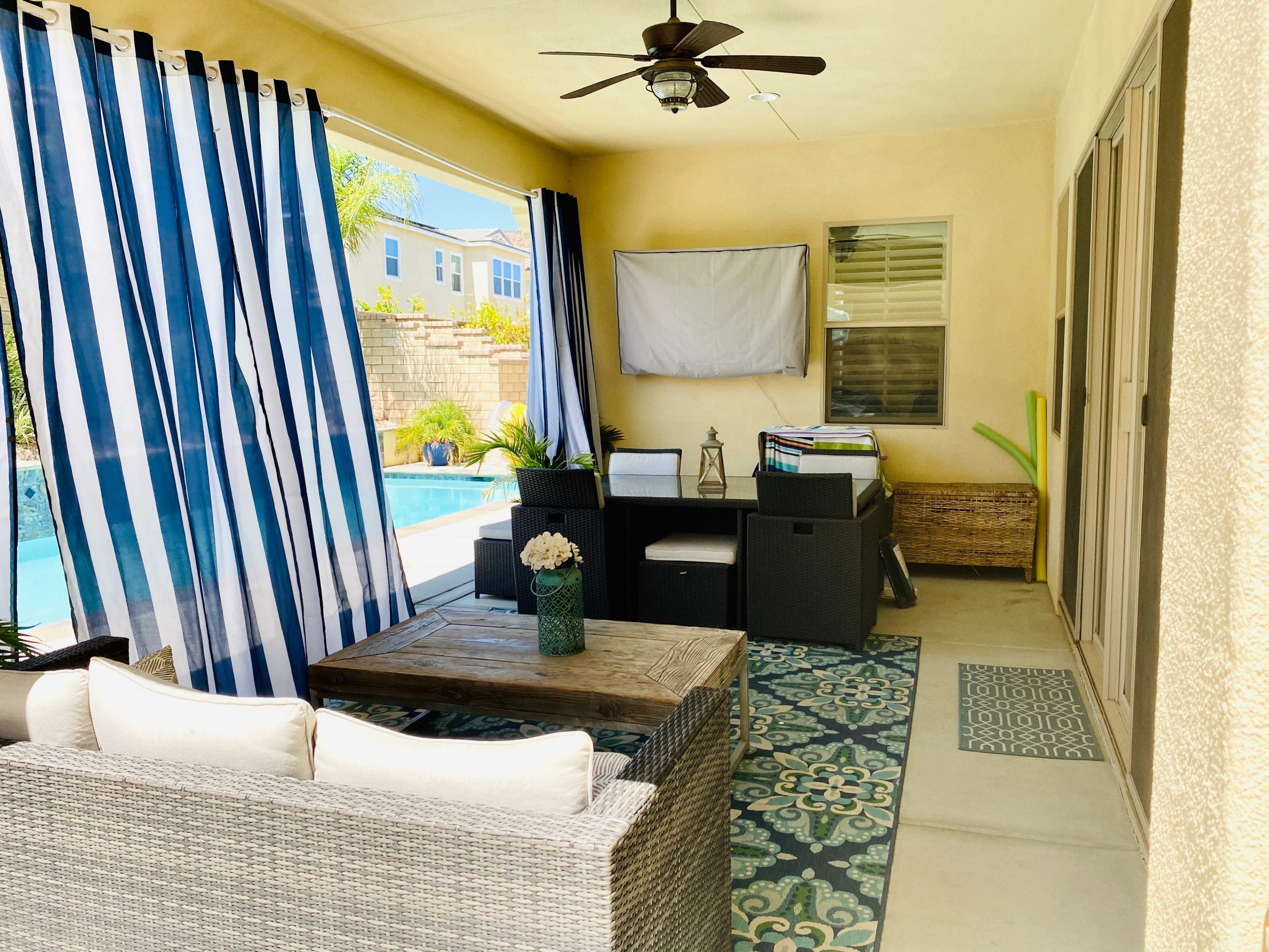 The image shows a covered patio area with striped curtains, a seating arrangement with a table, and a view of a swimming pool in the background.