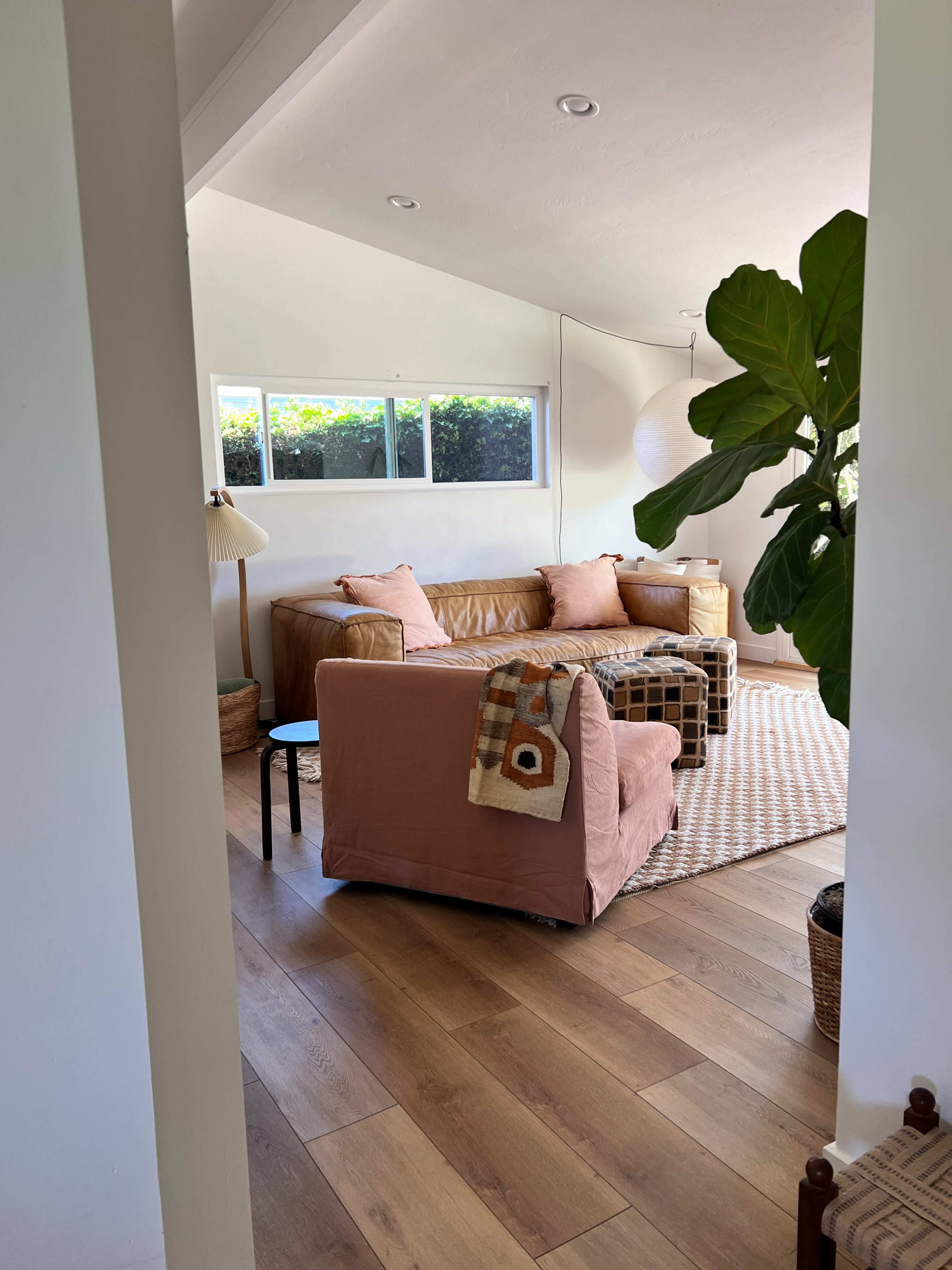 The image shows a cozy living room featuring a brown leather sofa, a pink upholstered chair, a coffee table, and a light-colored rug under natural light streaming in from large windows.